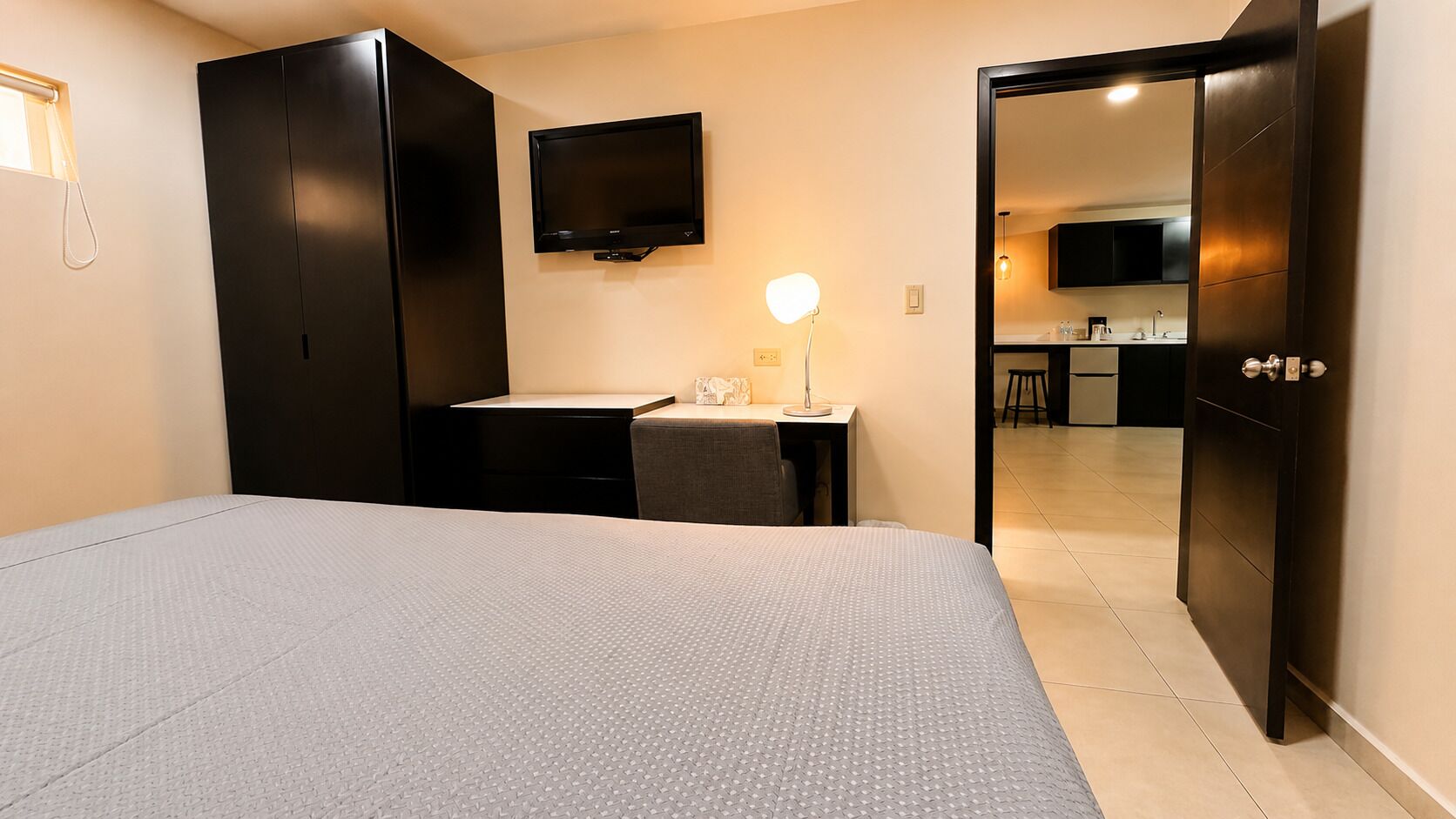 A hotel room with a bed, a desk with a lamp, a TV, and a kitchenette in the background.