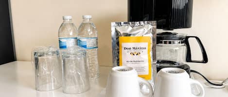 Una mesa con una cafetera, dos botellas de agua, un paquete de café y dos tazas blancas.