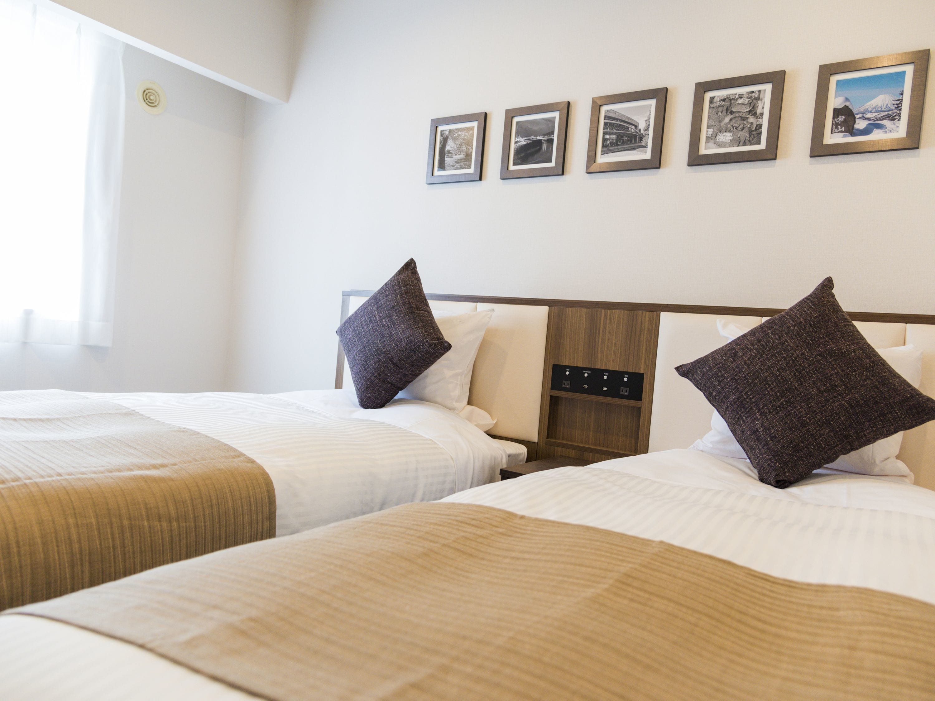 A hotel room with two beds, each with a wooden headboard, white bedding, and dark brown pillows. There are framed pictures on the wall above the beds.