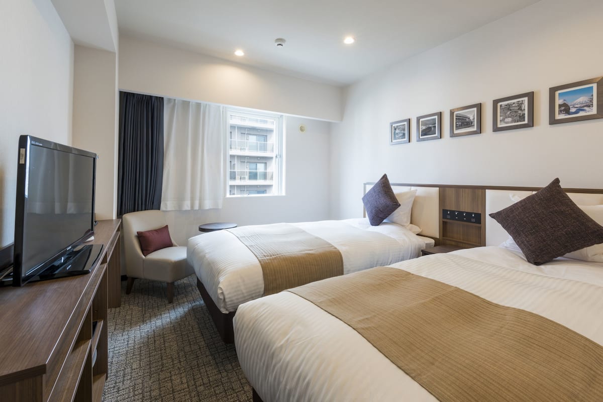 Ein Hotelzimmer mit zwei Betten, einem Fernseher, einem Schreibtisch und einem Fenster mit Vorhängen.