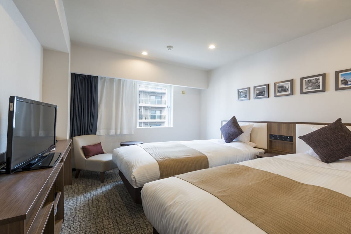 Ein Hotelzimmer mit zwei Betten, einem Fernseher, einem Schreibtisch und einem fensterbepflanzten Fenster.