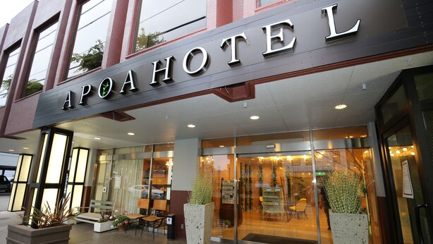 Exterior del hotel Apoi con fachada moderna, un amplio acceso de vidrio y un área de estar al aire libre con plantas en macetas.