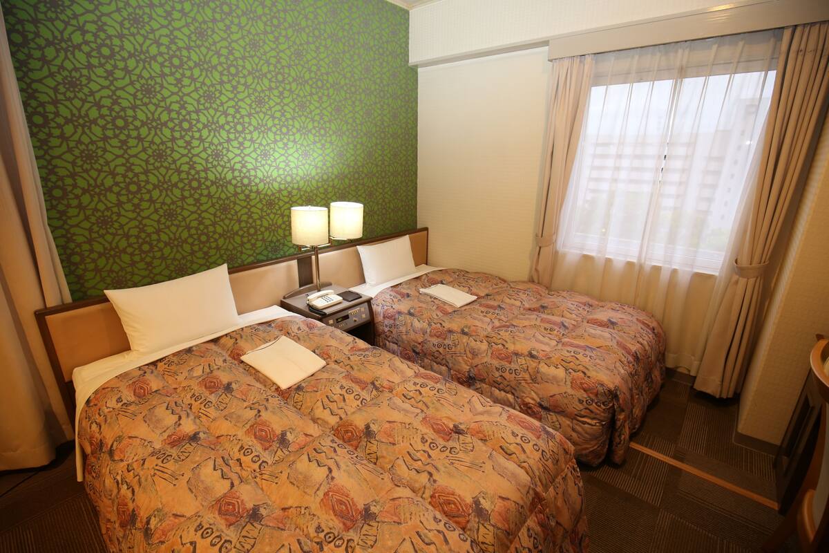 A hotel room with two beds, a green patterned wall, a window with curtains, and a nightstand with a lamp.