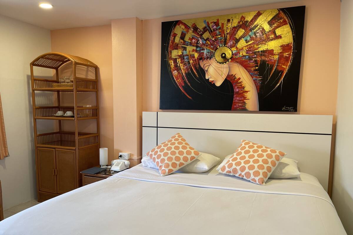 A hotel room with a bed, two patterned pillows, a nightstand with a phone, a lamp, a wooden shelf with glasses, and a painting on the wall.