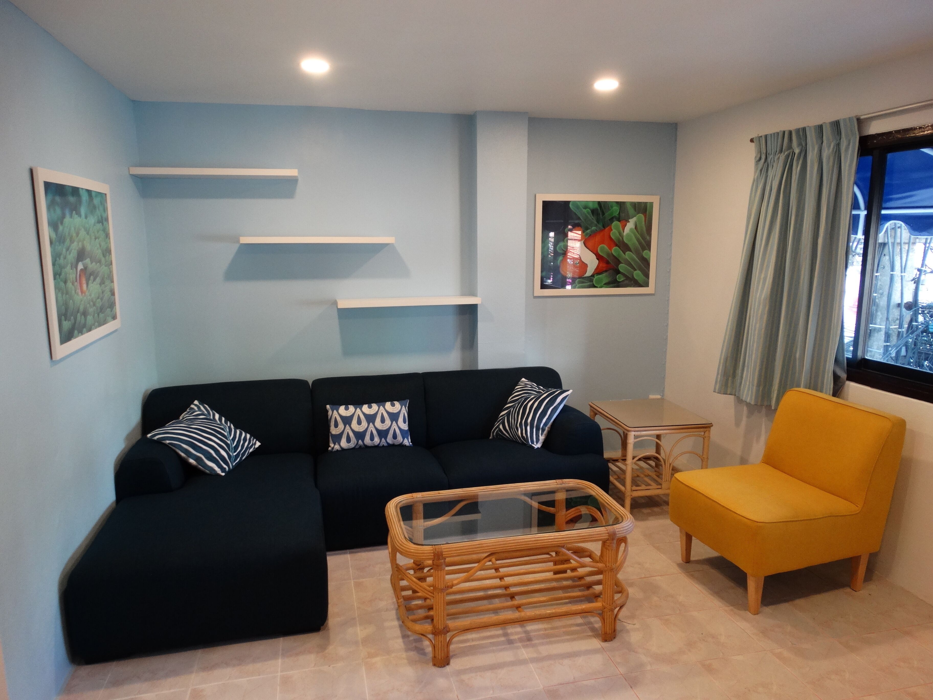 A living room with a black sofa, a yellow armchair, a glass-top coffee table, and a small side table. There are two wall-mounted shelves and two framed pictures.