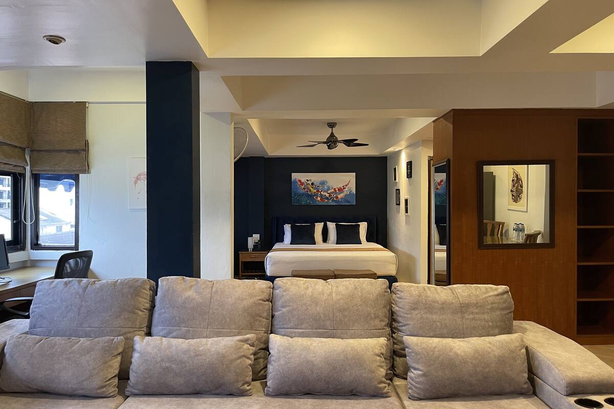 A modern hotel room with a large sofa, a bed with a dark headboard, a ceiling fan, and artwork on the walls.