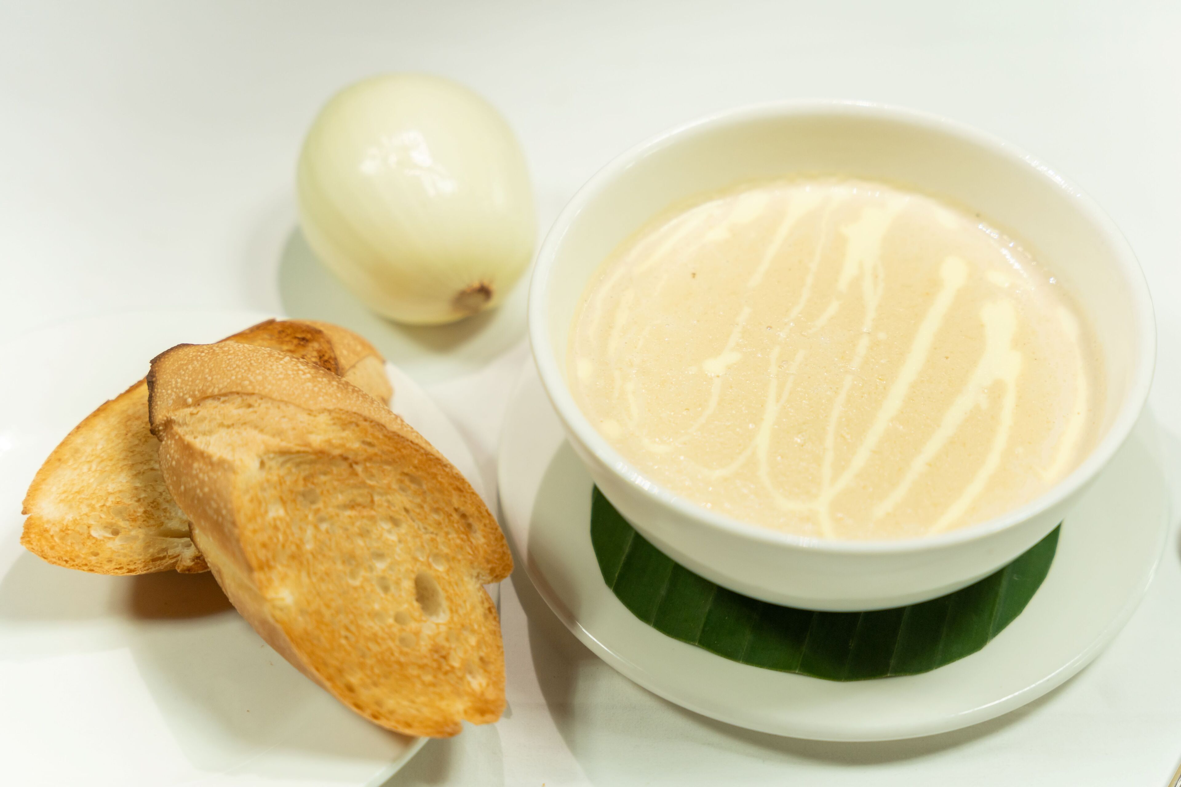 Un tazón de sopa con hoja de plátano, acompañado de dos tostadas y un huevo duro sobre un plato blanco.