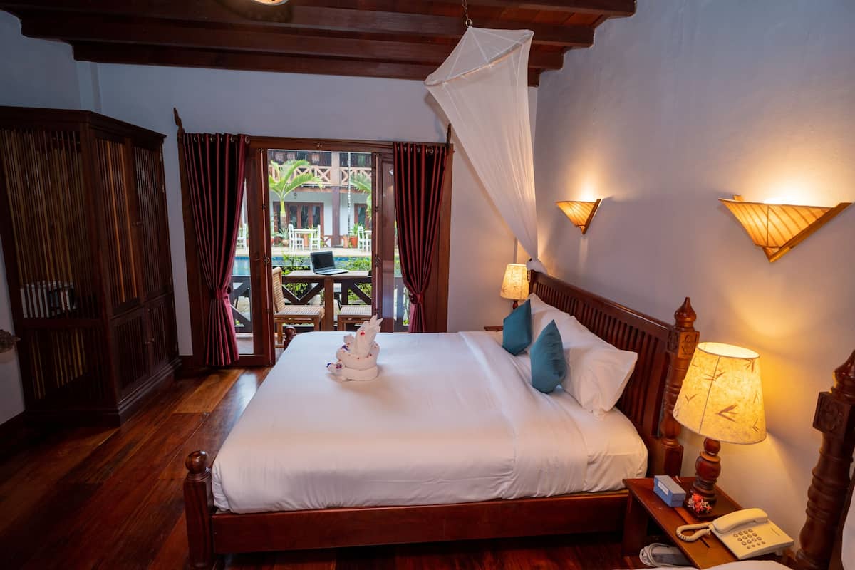 A hotel room with a wooden bed, a bedside table with a lamp, a telephone, and a view of an outdoor area with a table and chairs.