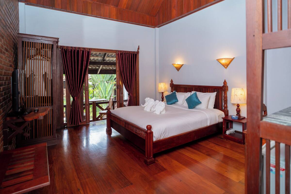 A spacious bedroom with a large wooden bed, two bedside lamps, and a view of greenery outside.