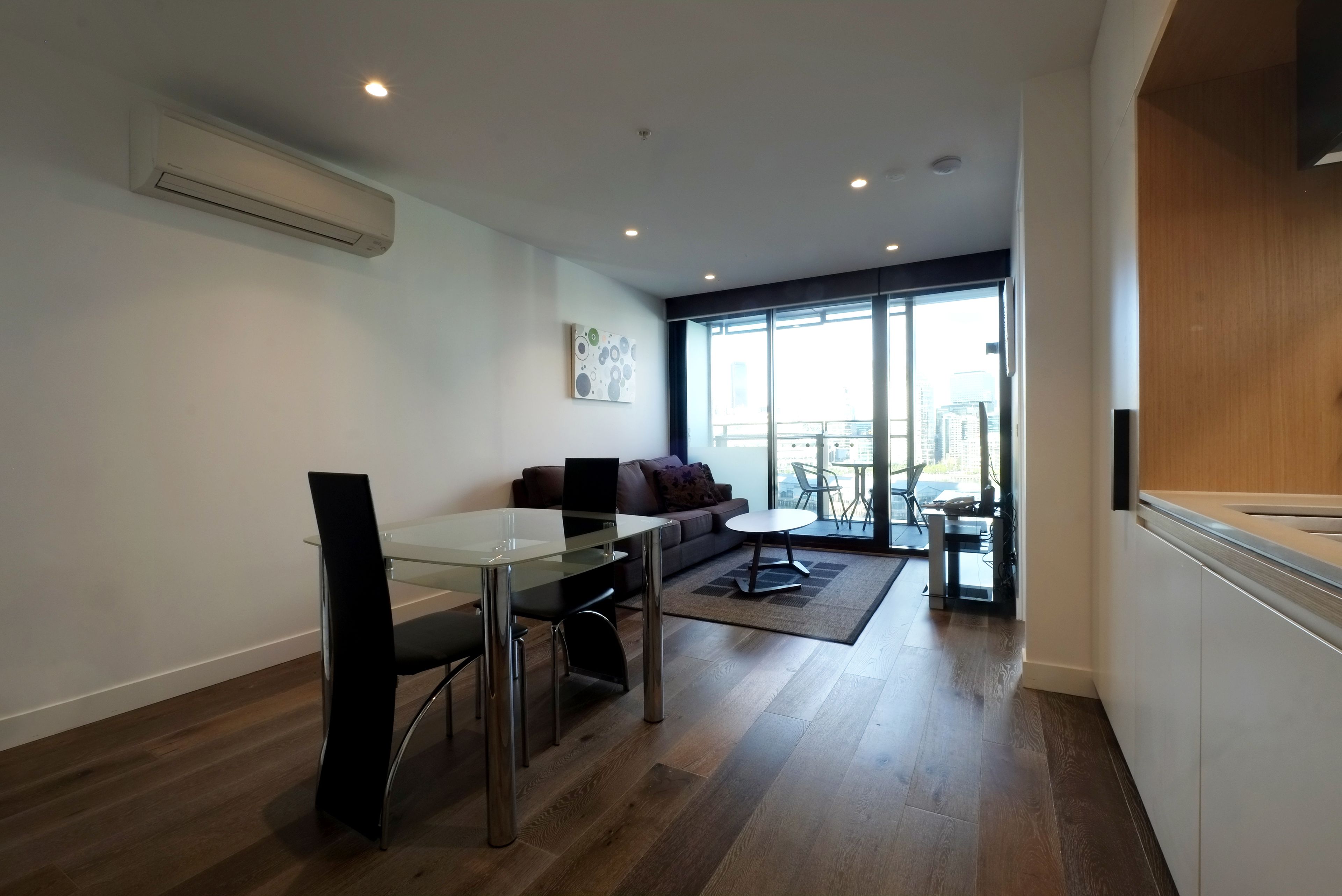A modern living room with a glass dining table, a sofa, and a view of the city through large windows.