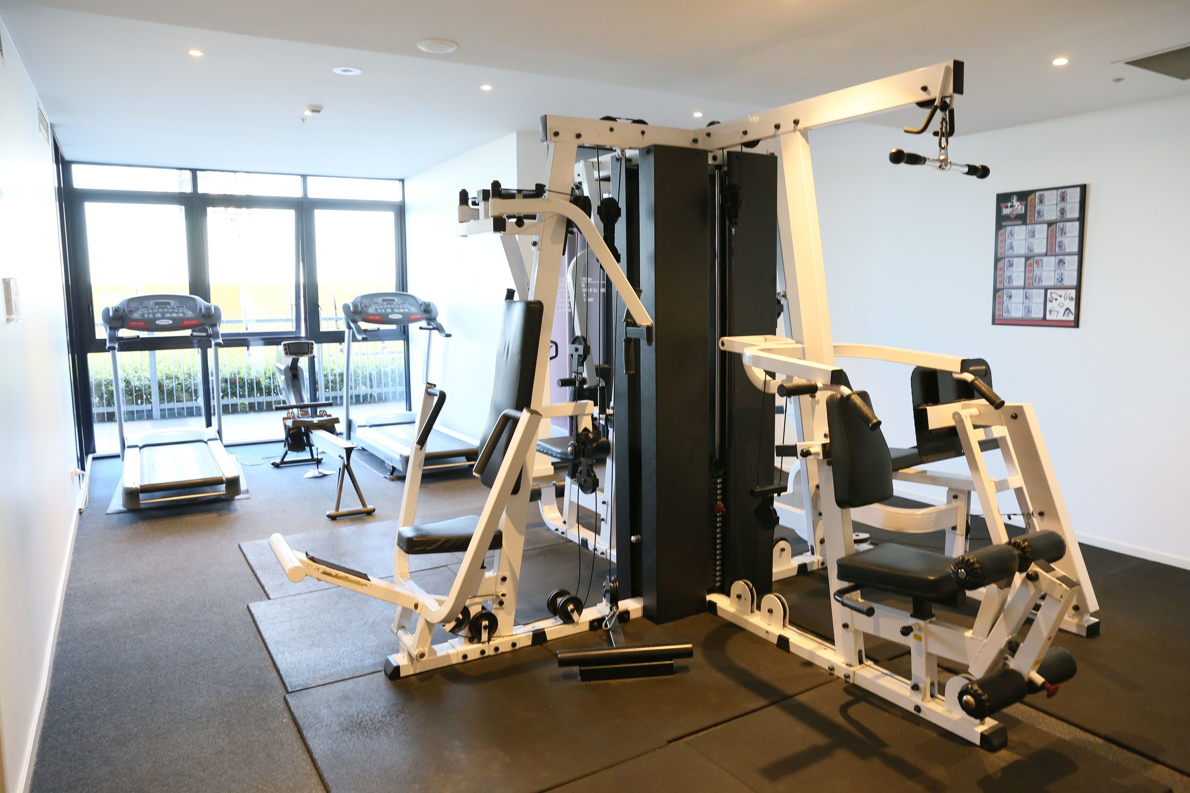 Ett gym med olika typer av tr äningsutrustning, ett löpband och ett stort fönster som släpper in naturligt ljus.