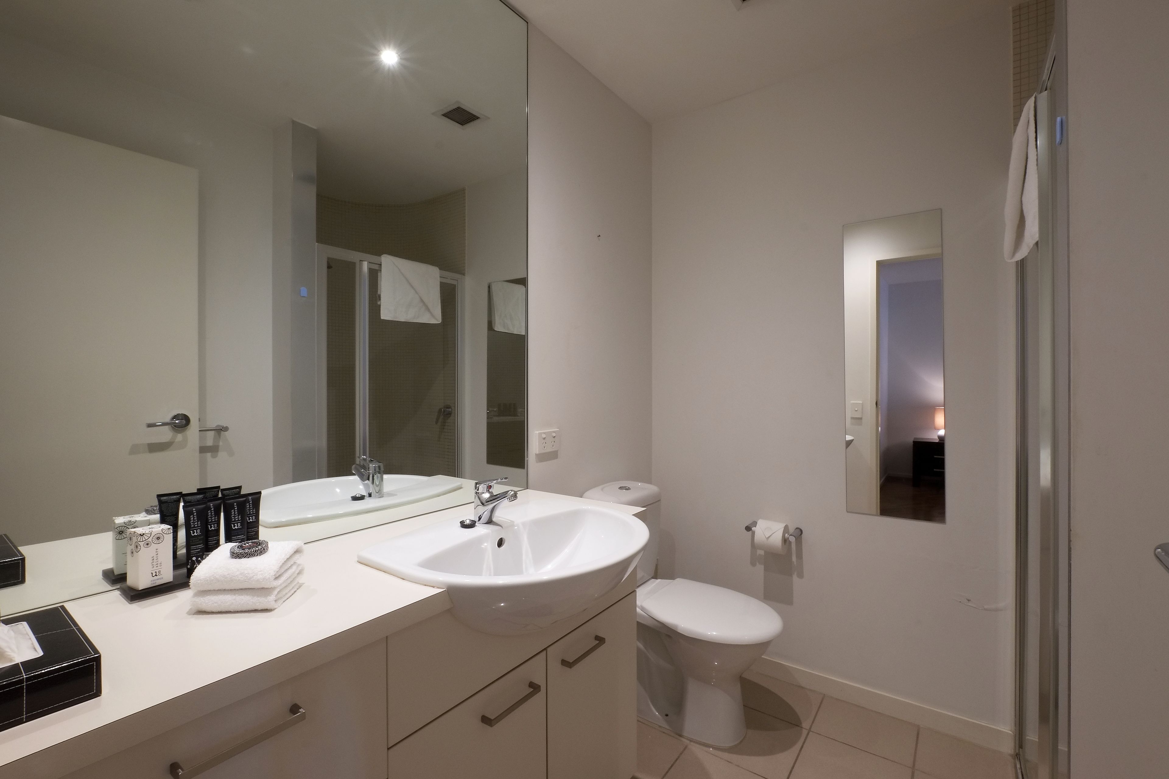 A modern bathroom with a large mirror, white sink, and toilet. There are towels and toiletries on the counter.