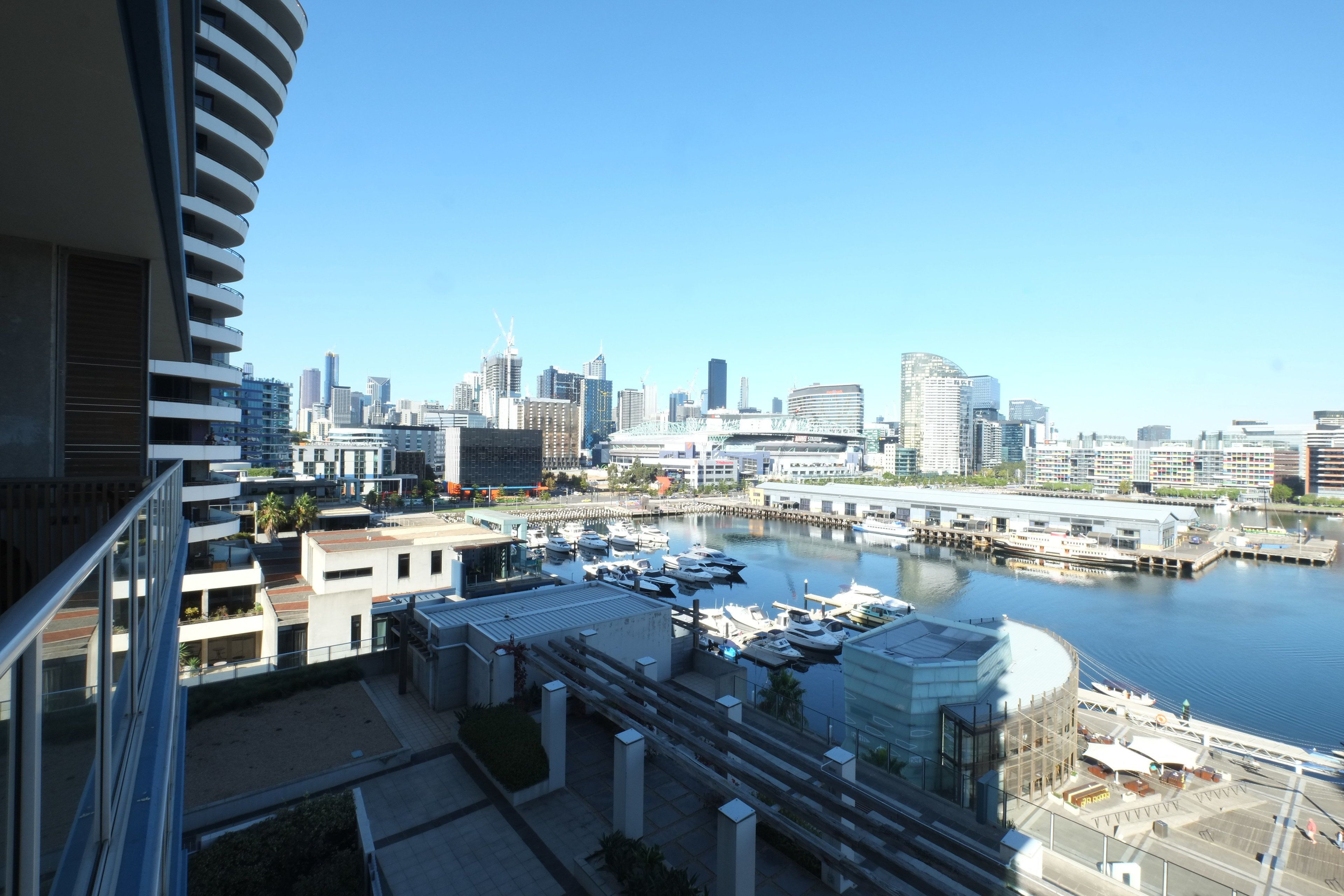 Uma marina com barcos atracados, um horizonte urbano ao fundo e edifícios modernos com varandas.