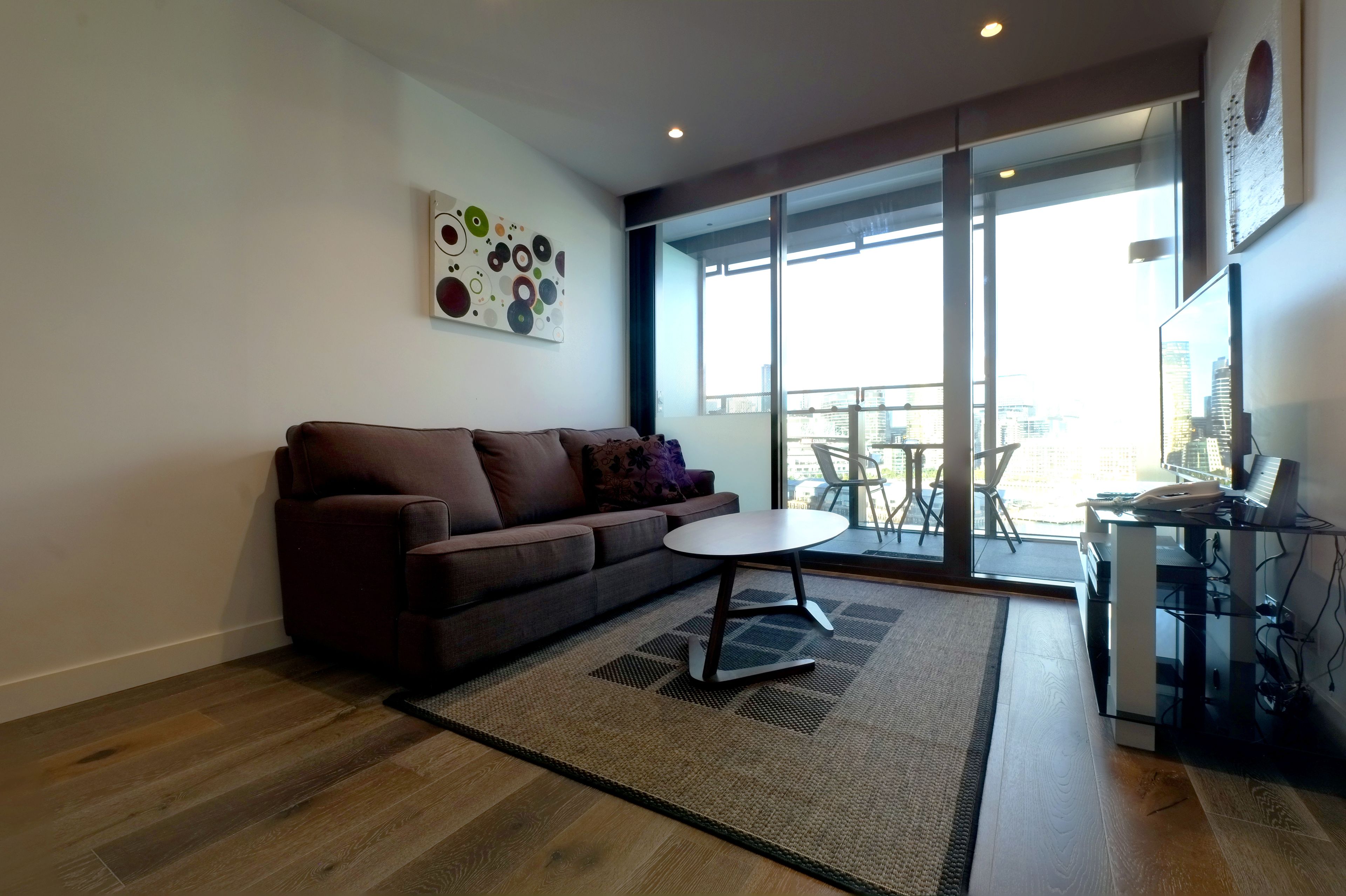 Ein modernes Wohnzimmer mit einem braunen Sofa, einem kleinen weißen Tisch und einem Glastisch mit einem Computer. Ein großes Fenster bietet Blick auf die Stadt und an der Wand hängt ein Gemälde.