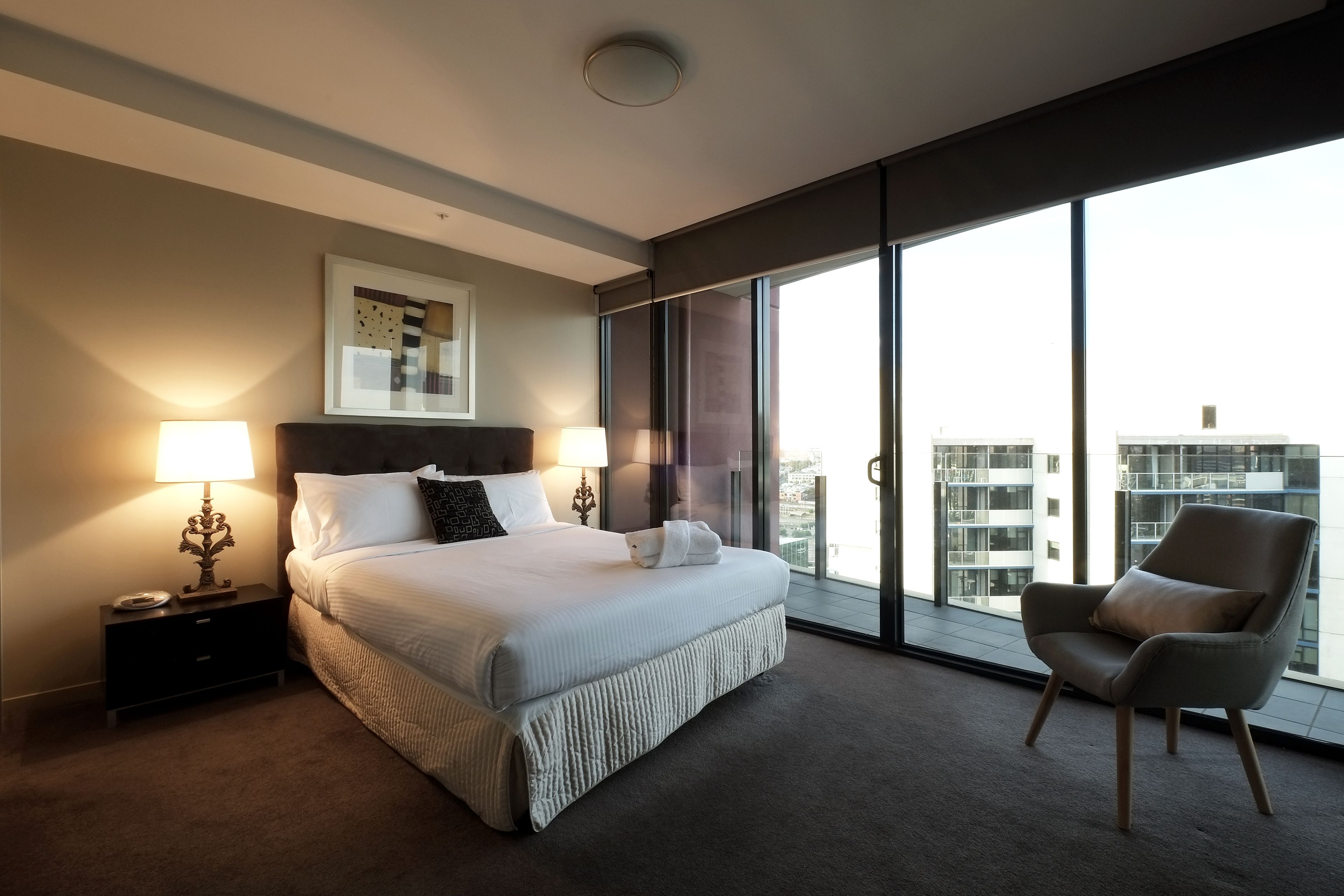A modern hotel room with a large bed, two bedside lamps, a chair, and a large window with a city view.