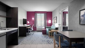 A modern hotel room with a kitchen area, a dining table with chairs, a desk with a computer, and a large window with curtains.