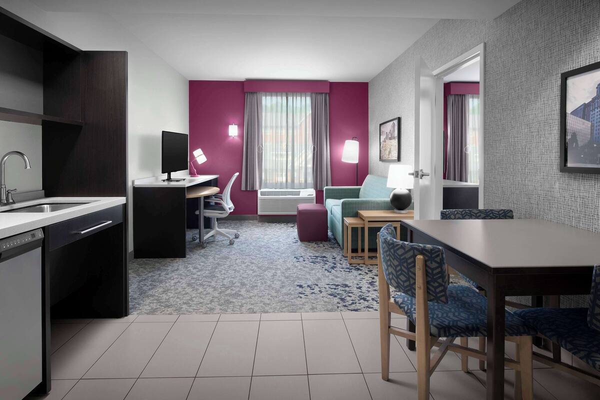 A modern hotel room with a kitchen area, a dining table with chairs, a desk with a computer, and a large window with curtains.