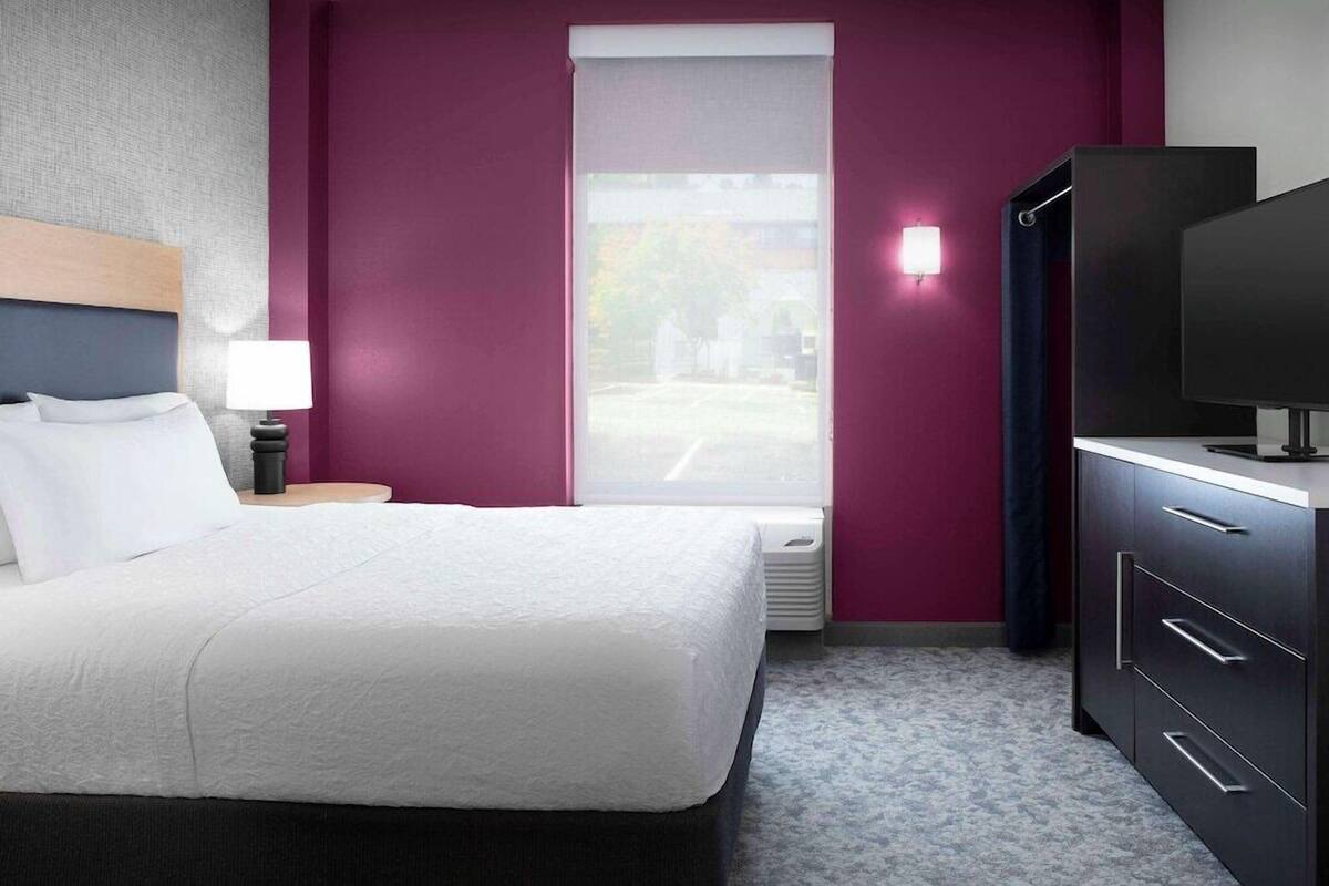 A hotel room with a bed, a TV, a nightstand, and a window with a view of the outside.