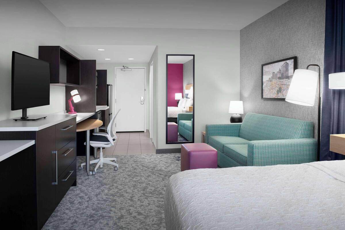 A modern hotel room with a bed, desk, chair, and a large mirror.