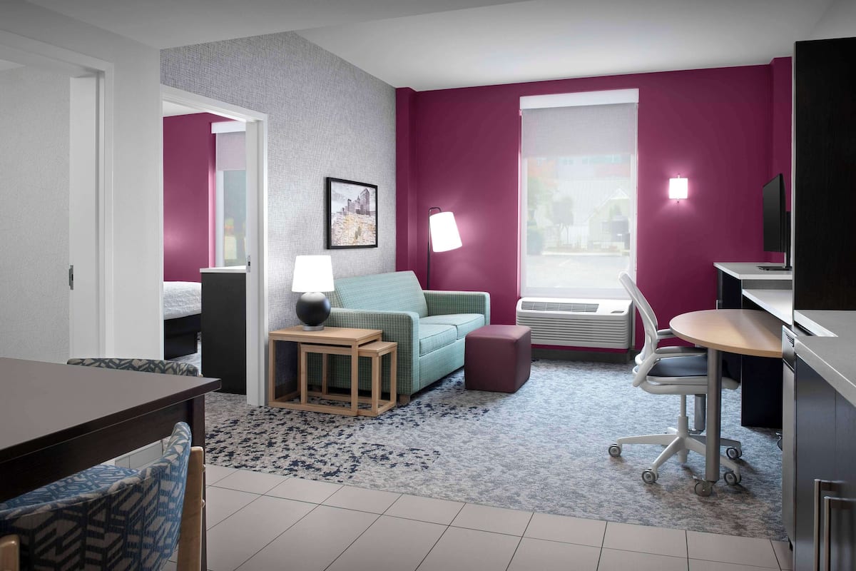 A modern hotel room with a teal sofa, a wooden side table, a desk with a chair, and a large window with a view.