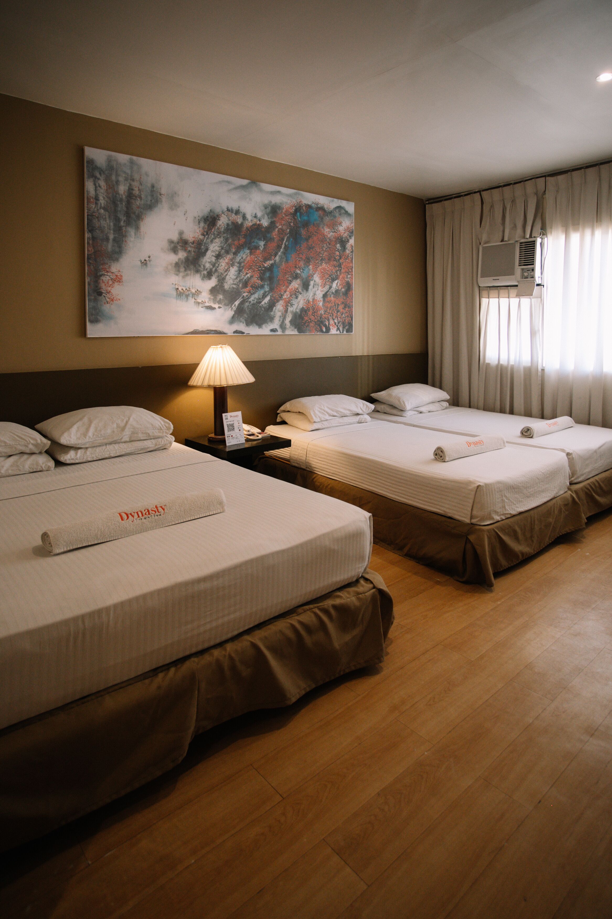 Habitación de hotel con tres camas, un cuadro en la pared, un equipo de aire acondicionado y una lámpara en la mesita de noche.