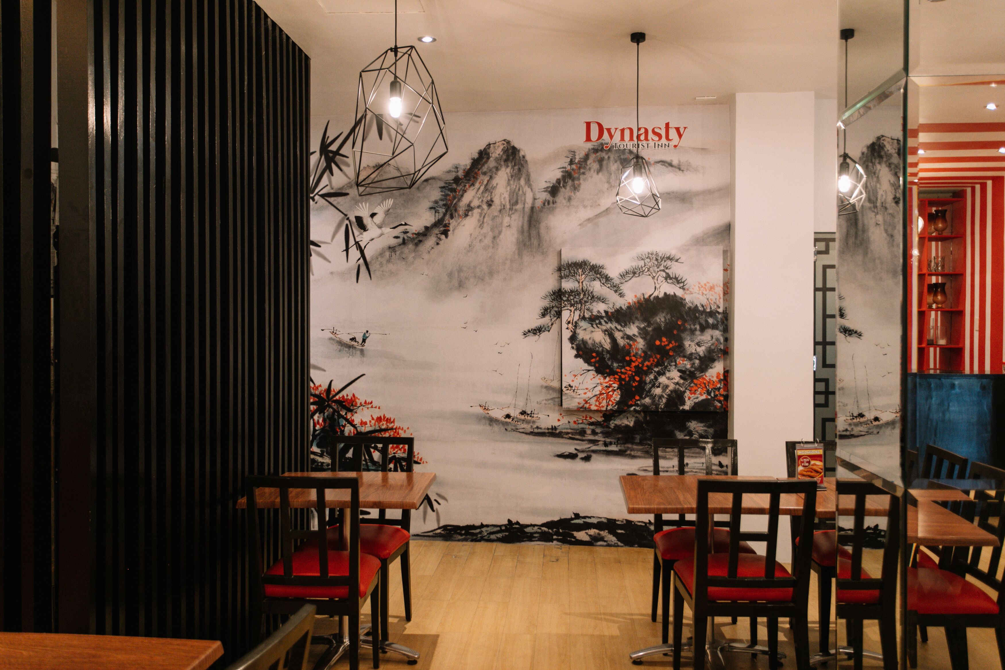 A restaurant with wooden tables and red chairs, a mural with mountains and birds, and hanging geometric light fixtures.