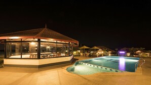 Un área de piscina con un salón envuelto en cristal, una terraza con baldosas y una pequeña cascada.