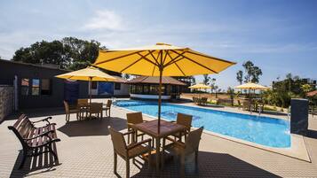 Zona junto a la piscina con solárium de baldosas, sillas reclinables de madera y sombrillas amarillas.
