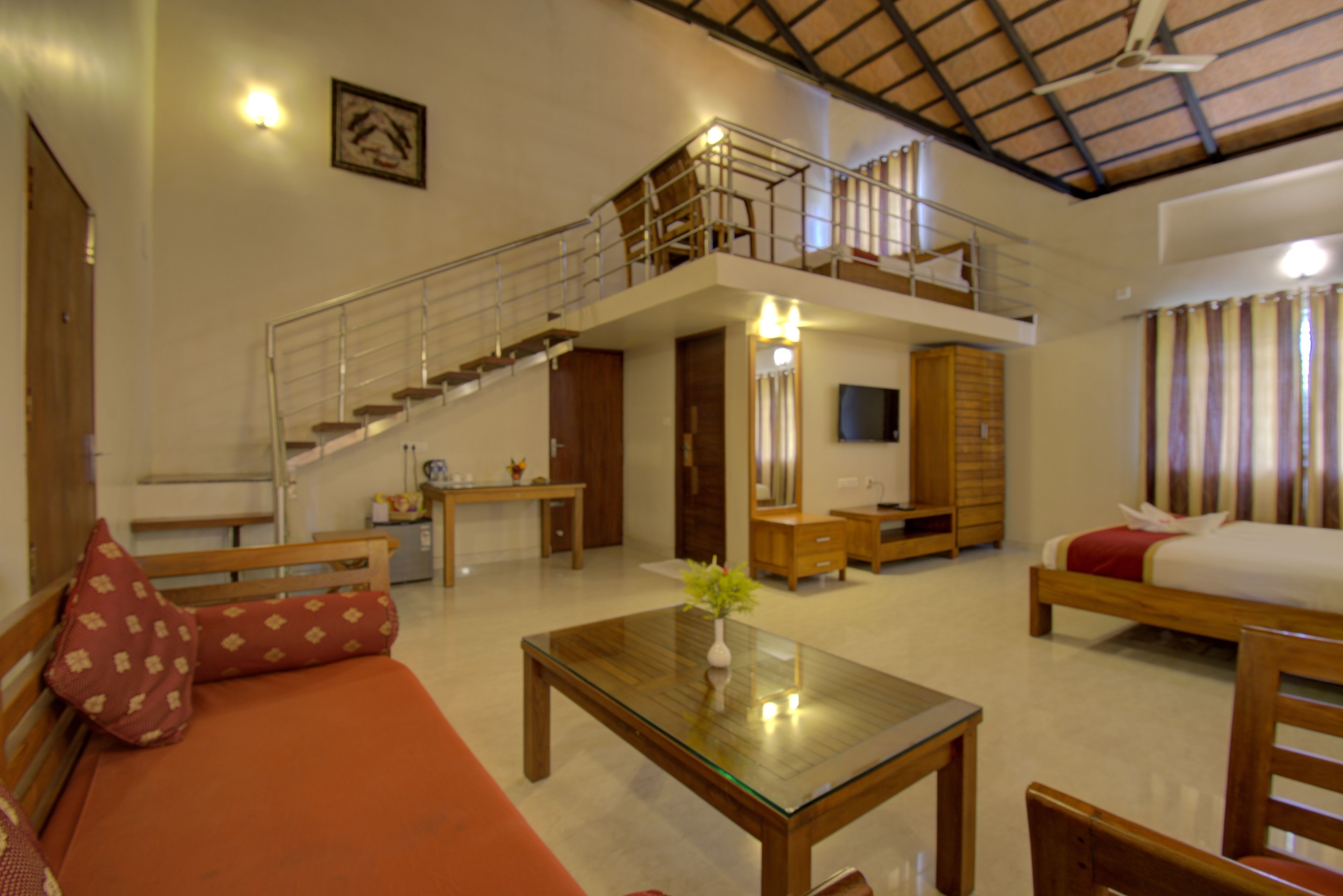 A spacious living area with a sofa, a glass-top coffee table, a staircase leading to an upper level, and a bed with a red blanket.