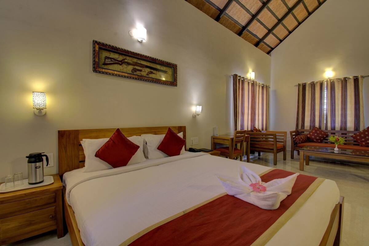 Habitación de hotel con una cama grande, cabecera de madera, una mesa de comedor con sillas, un sofá y un cuadro colgado en la pared.