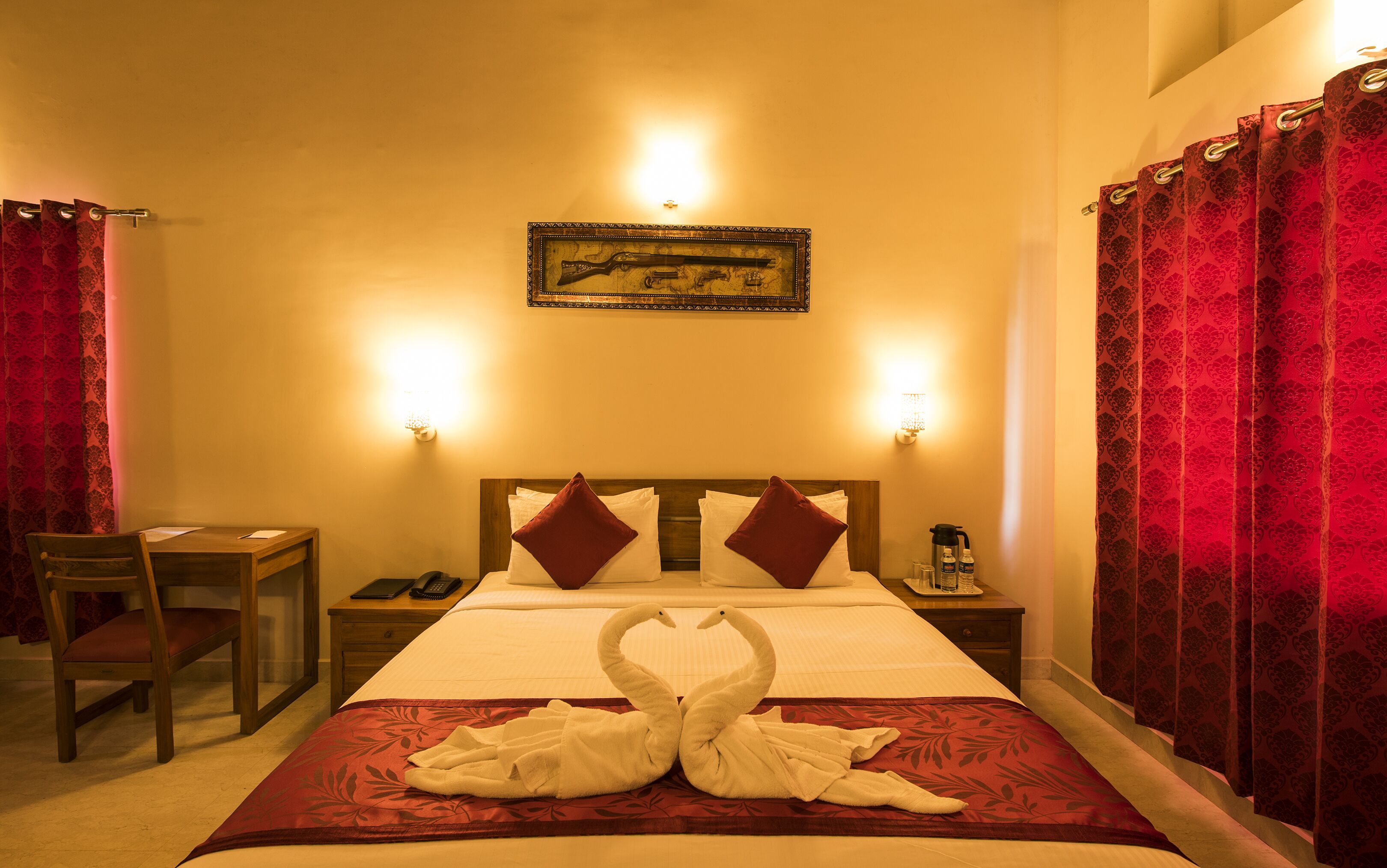 A hotel room with a bed, two bedside lamps, a wooden nightstand, a desk, a chair, and a wall-mounted picture frame.