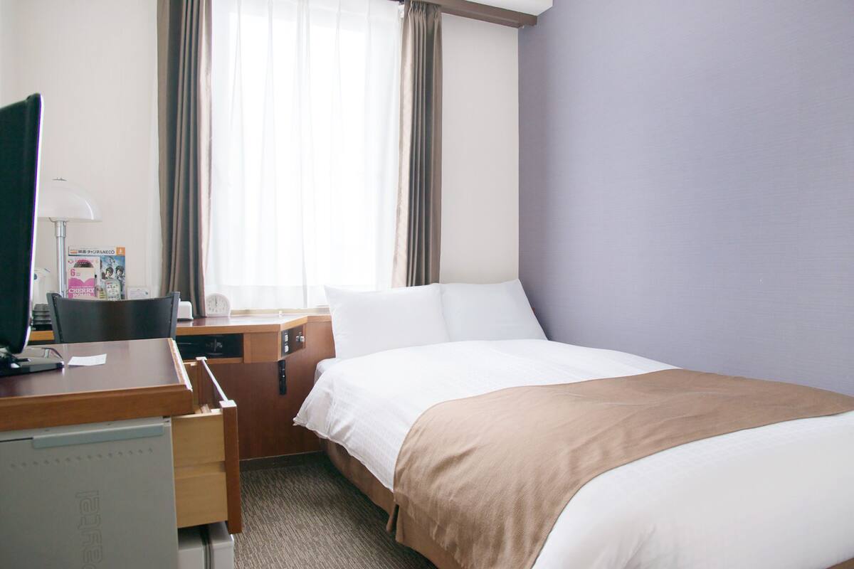 Une chambre d’hôtel avec un lit, un bureau avec un ordinateur, une chaise et une fenêtre avec des rideaux.