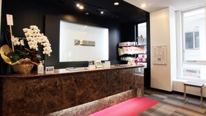 A hotel reception desk with a marble counter, a digital screen displaying the hotel name, a potted orchid, and a red carpet leading to the entrance. - Hotel Abest Nagano Ekimae (Nagano)