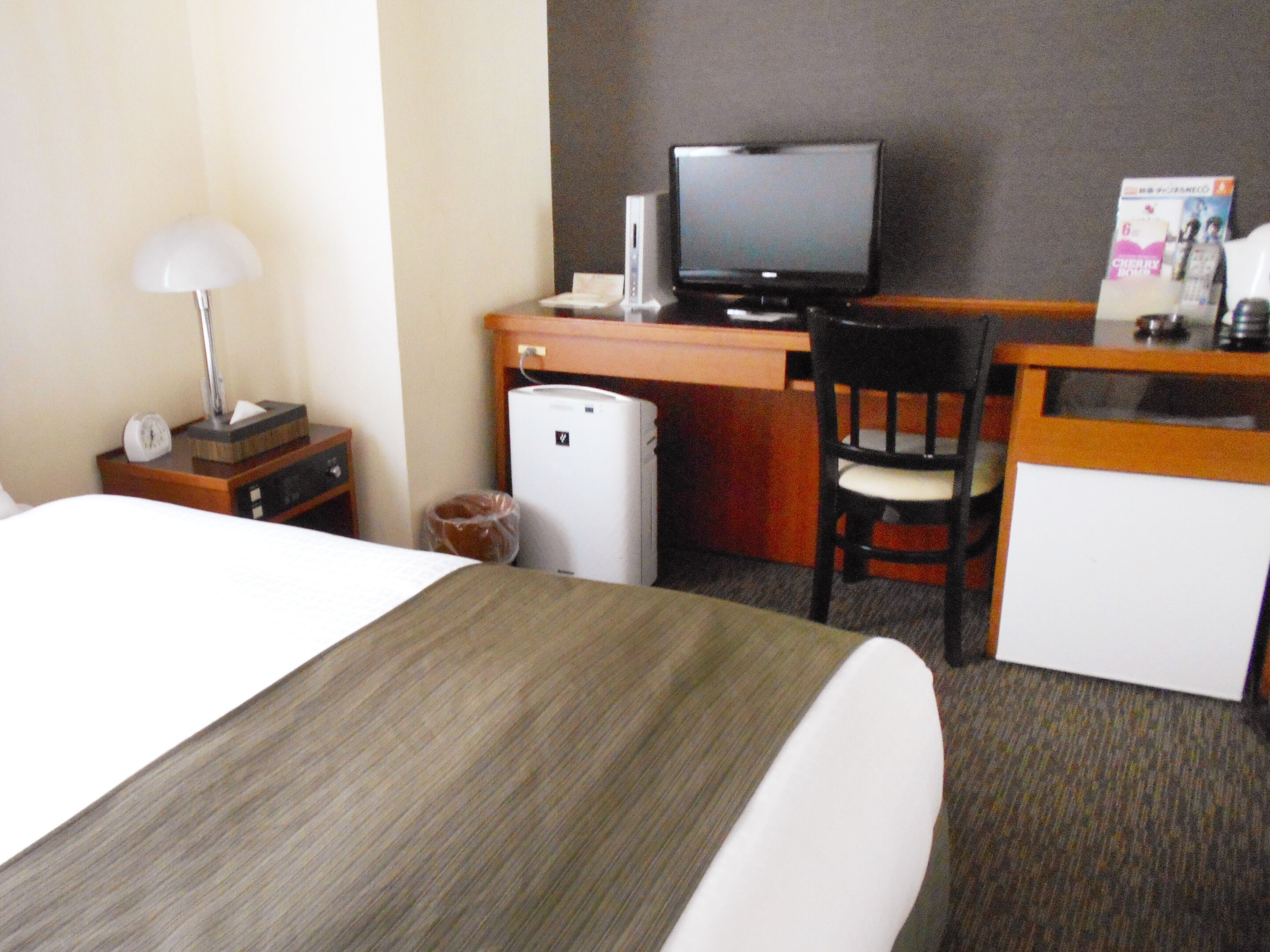 A hotel room with a bed, a desk with a computer, a chair, a small refrigerator, and a lamp.