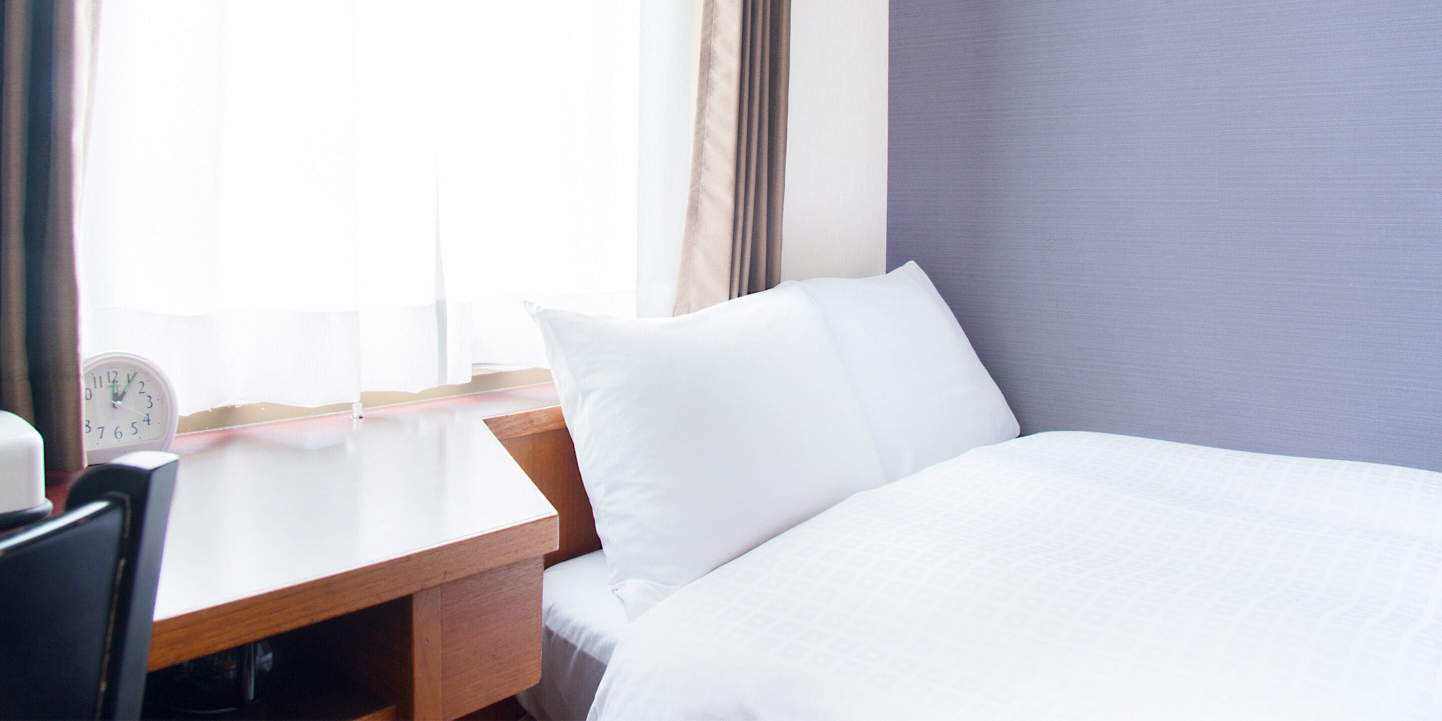 A hotel room with a bed, a wooden nightstand, a white pillow, a white bedspread, a clock, and a window with curtains.