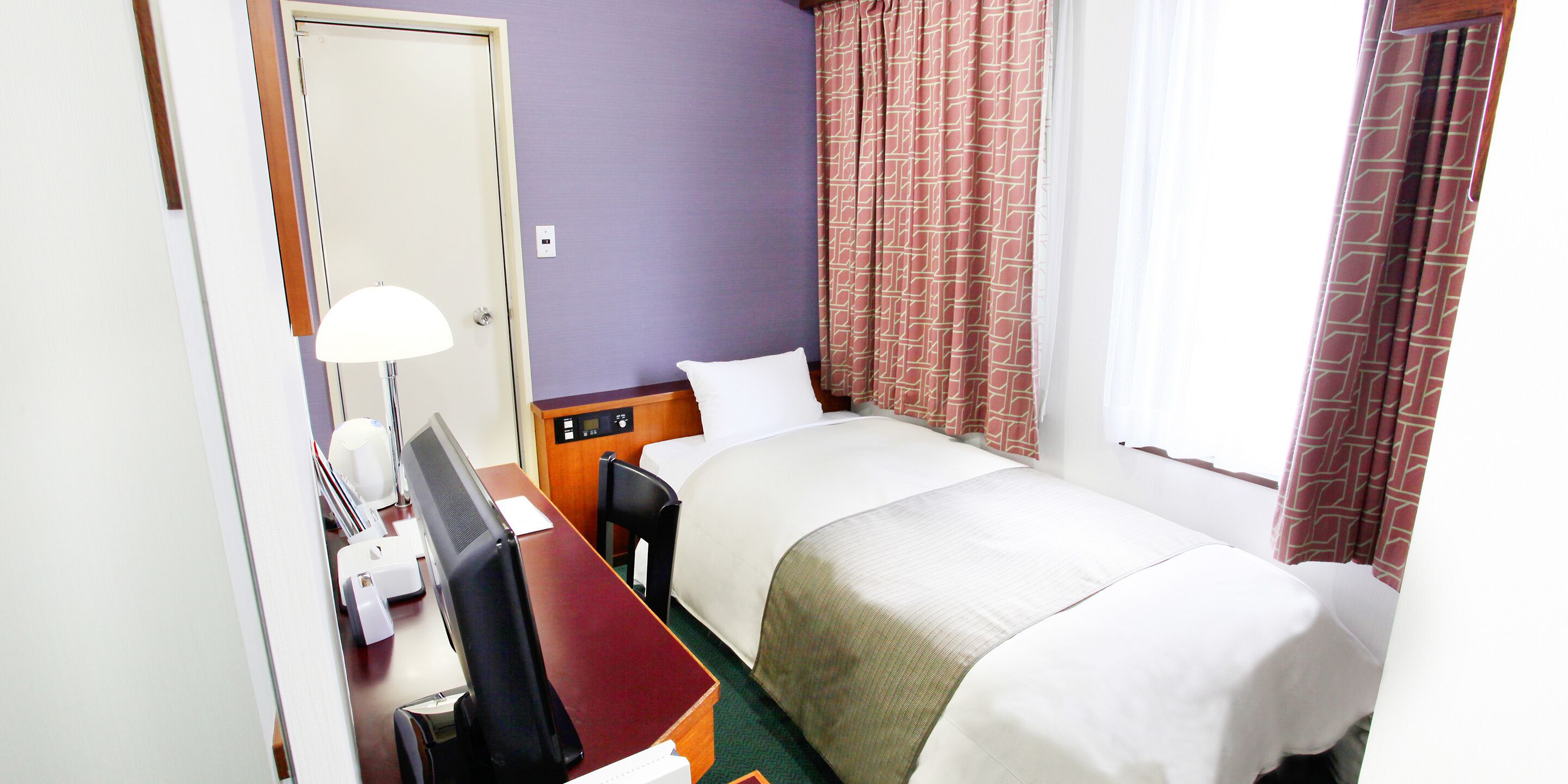 A hotel room with a bed, a desk with a computer, a chair, and a lamp.