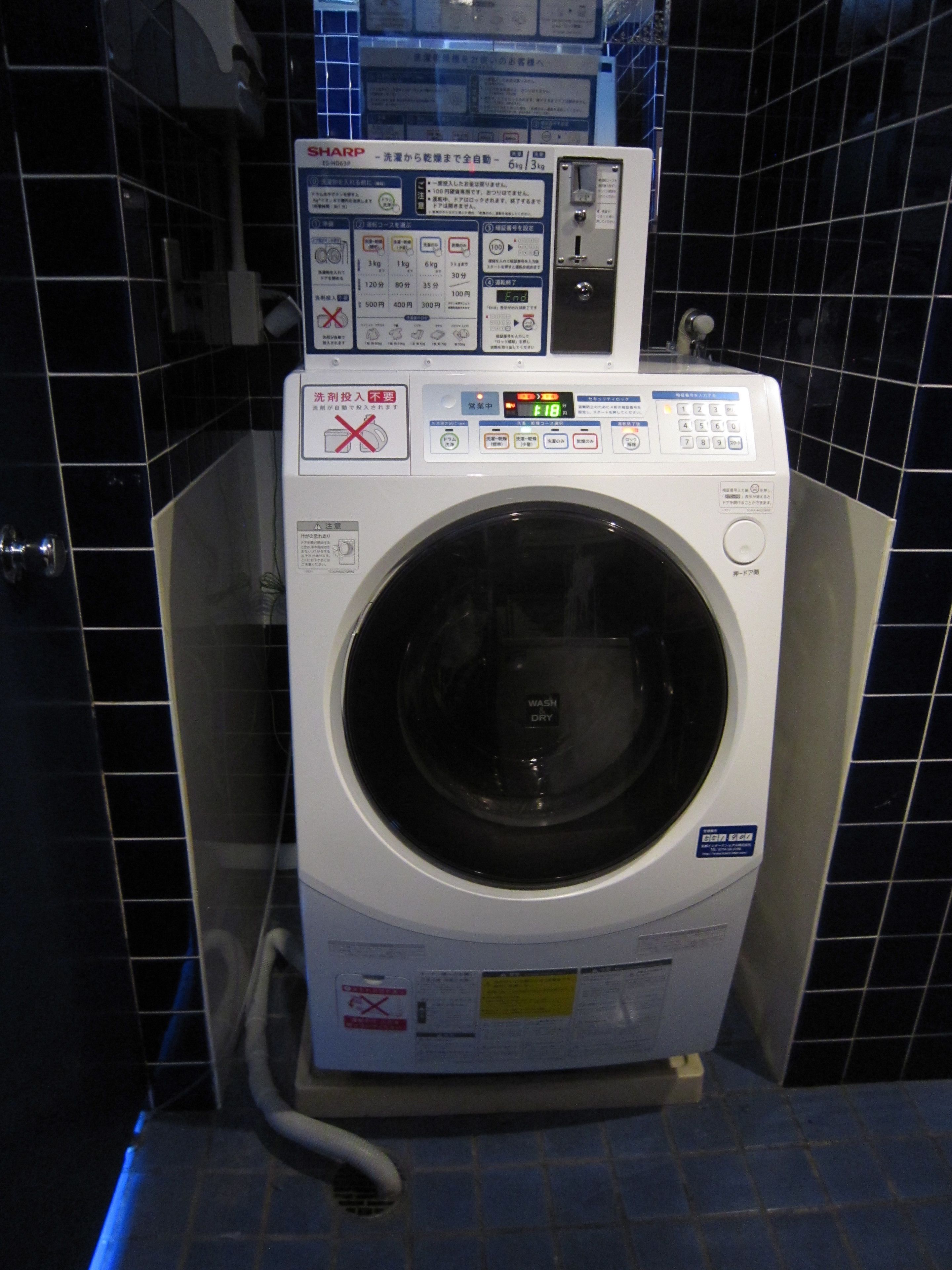 A front-loading washing machine with a control panel, located in a room with black tiled walls.
