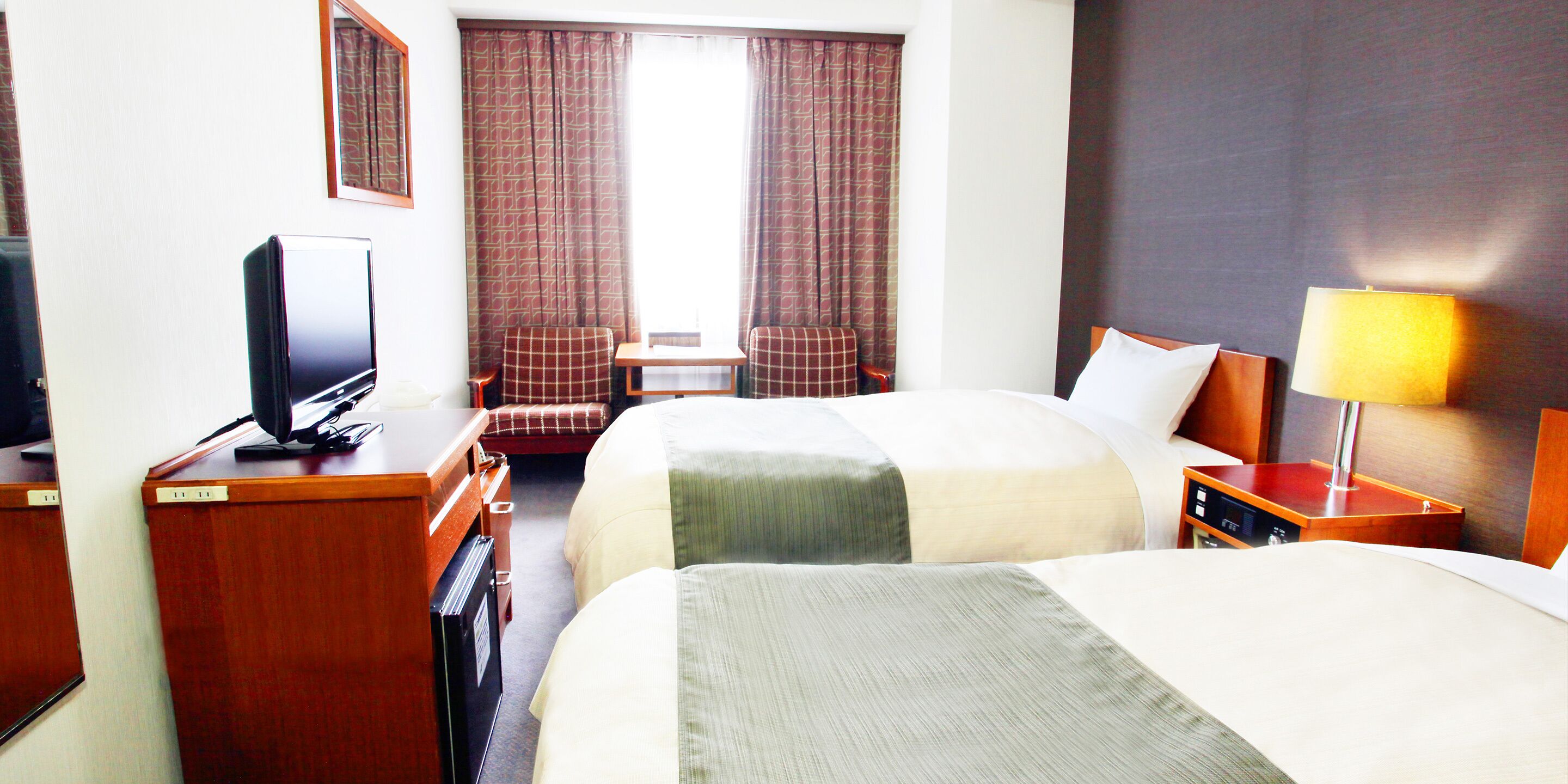 Habitación de hotel con dos camas, un escritorio, televisor, una silla, lámpara y una ventana con cortinas.