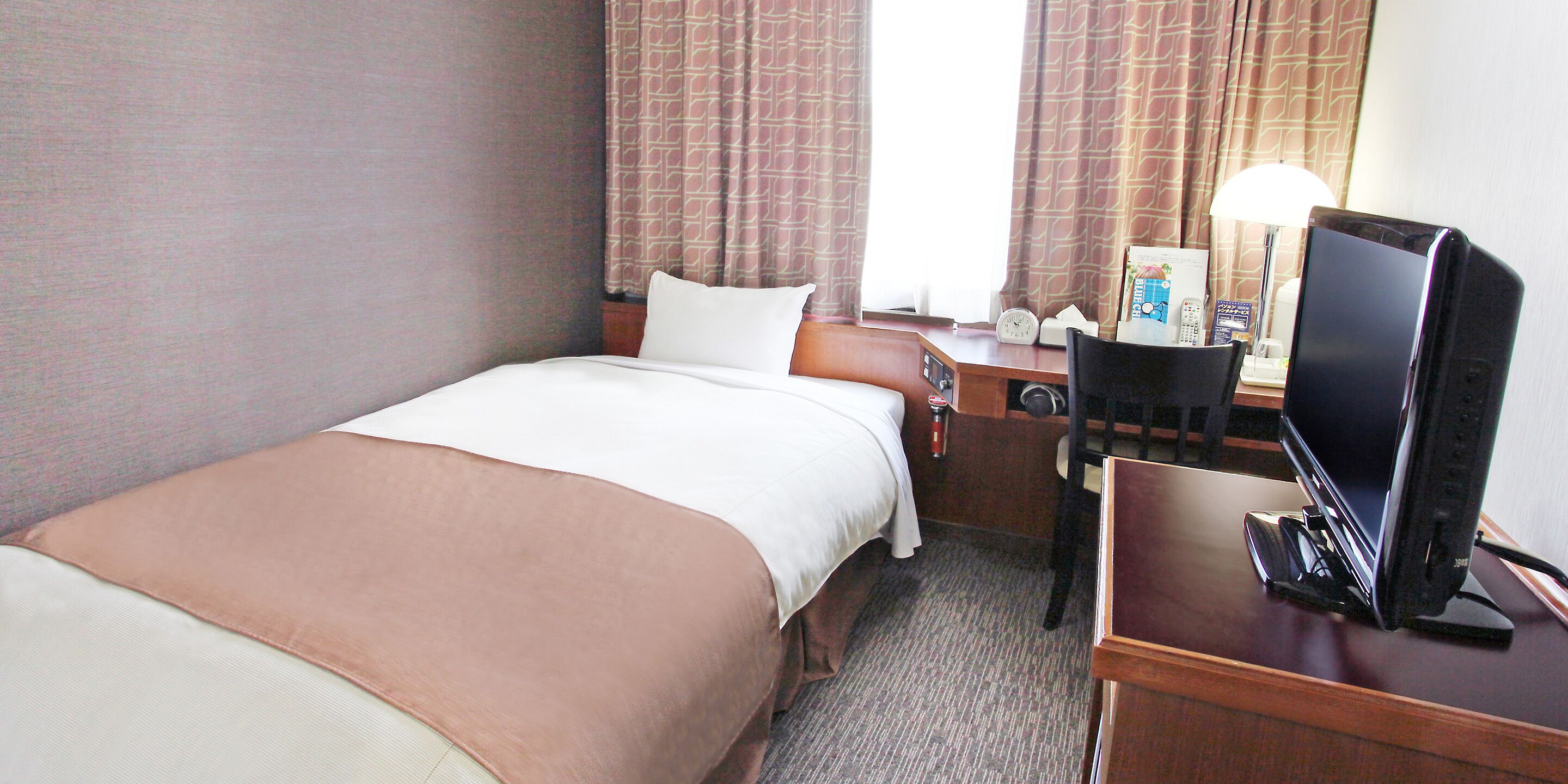 A hotel room with a bed, a desk with a computer, a chair, a lamp, and a clock.