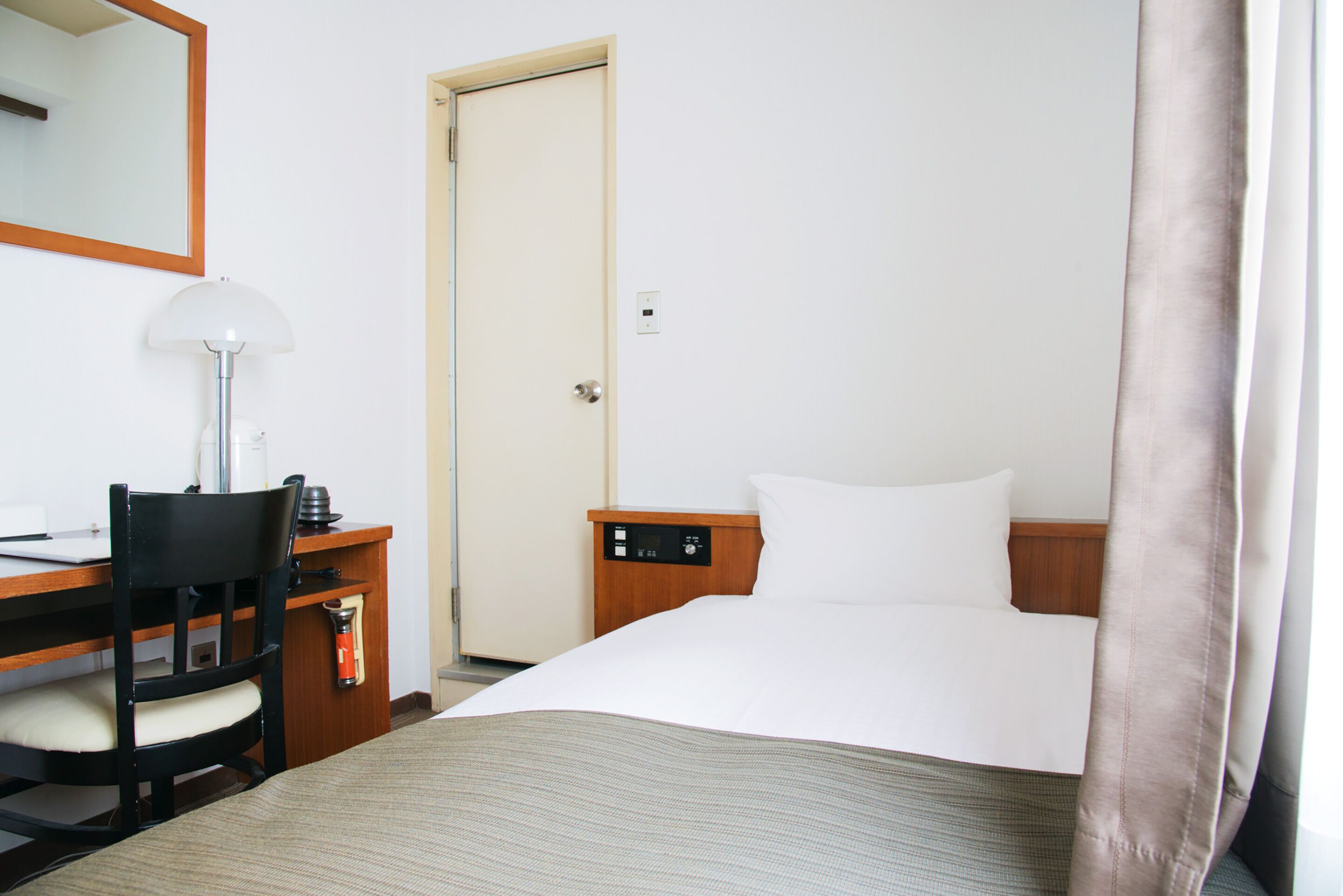 Una habitación de hotel con cama, escritorio, silla, lámpara, mesita de noche y puerta.