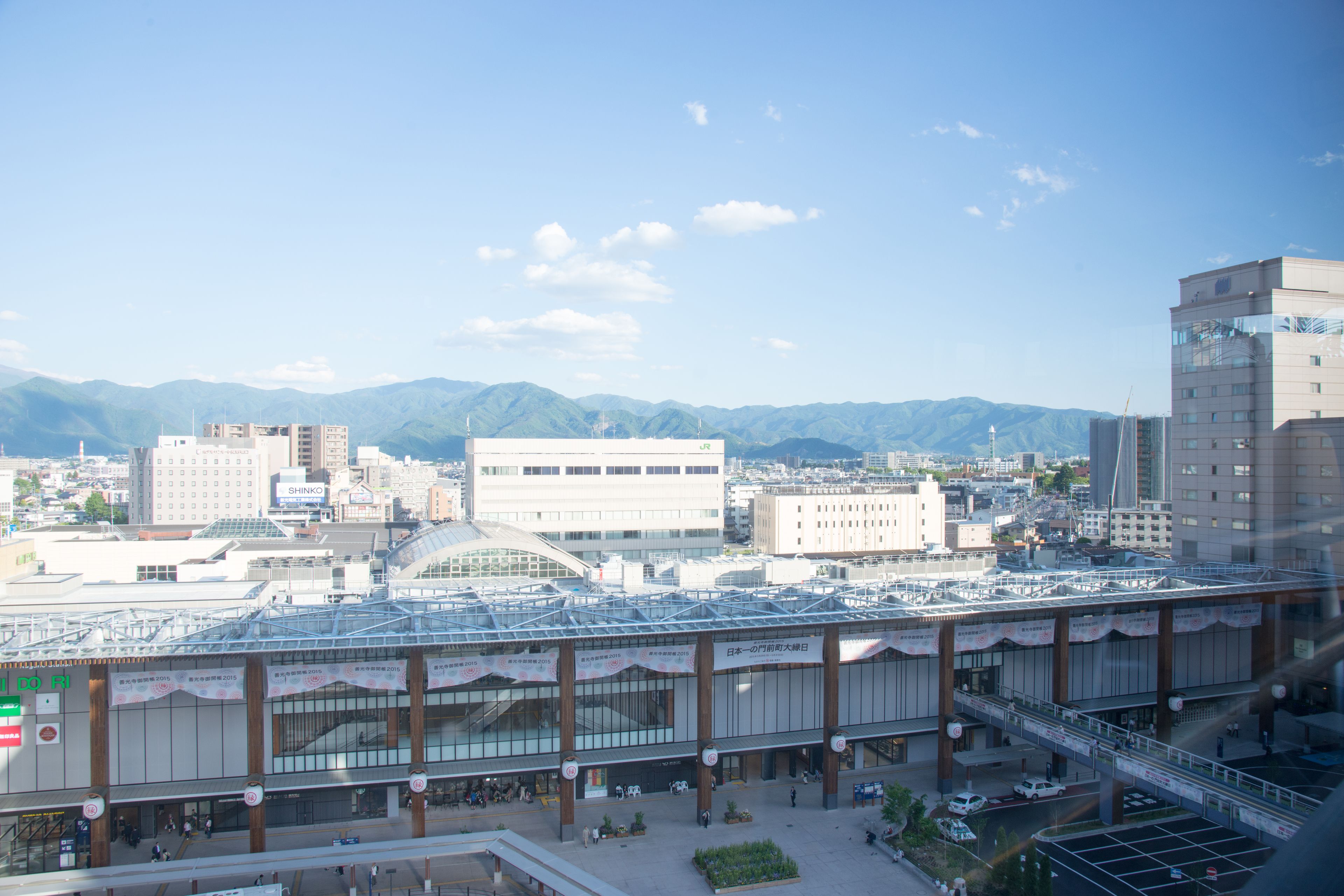 A train station with a modern glass roof, surrounded by urban buildings and mountains in the background.