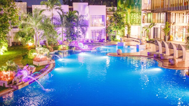 Un área de piscina nocturna con varias piscinas, palmeras y edificios modernos.