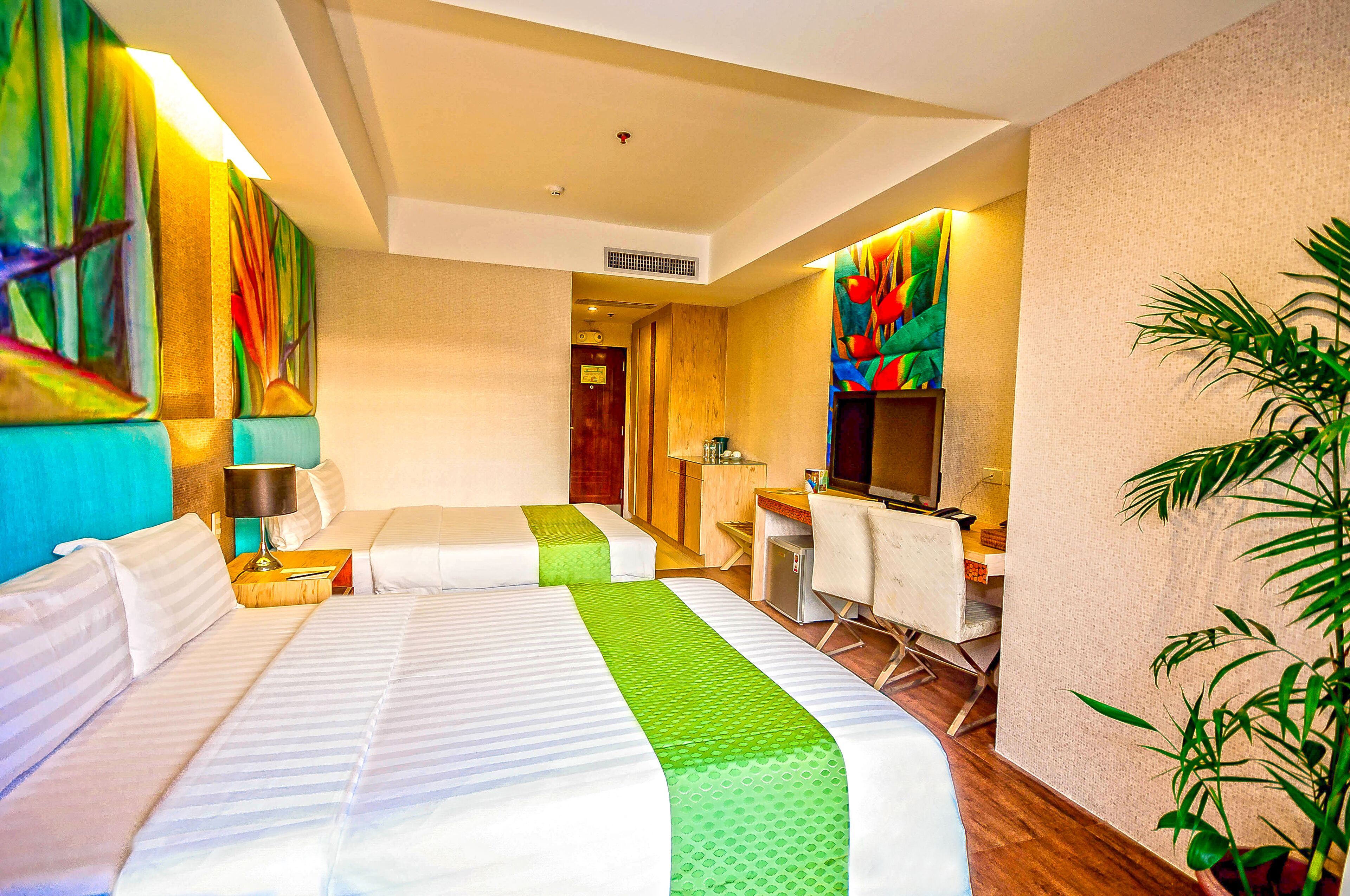A hotel room with two beds, a desk, a chair, a television, and a plant.