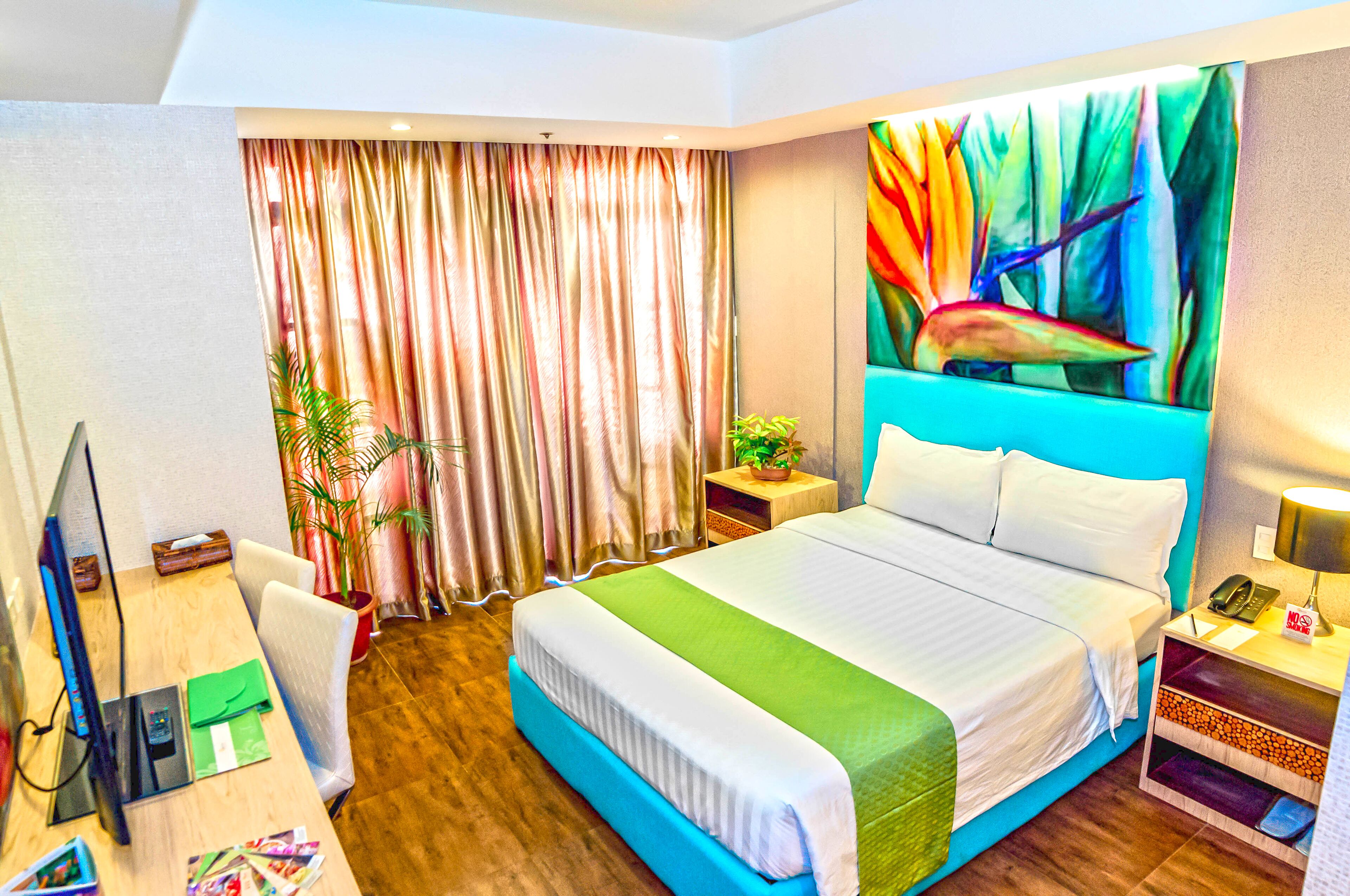 A hotel room with a large bed, a desk with a computer, a chair, a plant, and a painting on the wall.