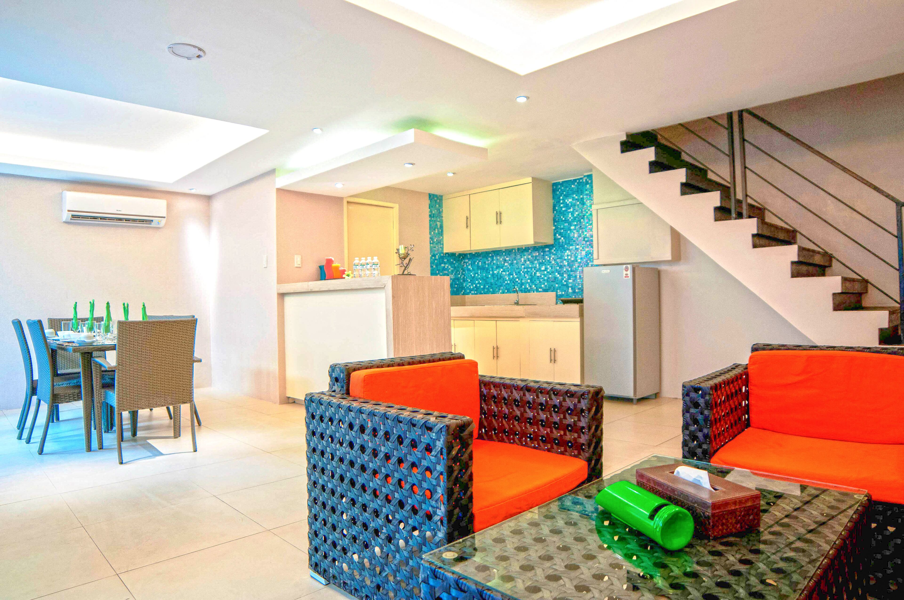 A modern living area with a dining table, chairs, and a sofa. There is a kitchen in the background with cabinets and a refrigerator.