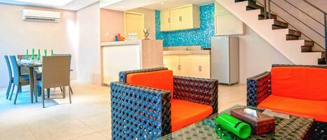A modern living area with a dining table, chairs, and a sofa. There is a kitchen in the background with cabinets and a refrigerator.
