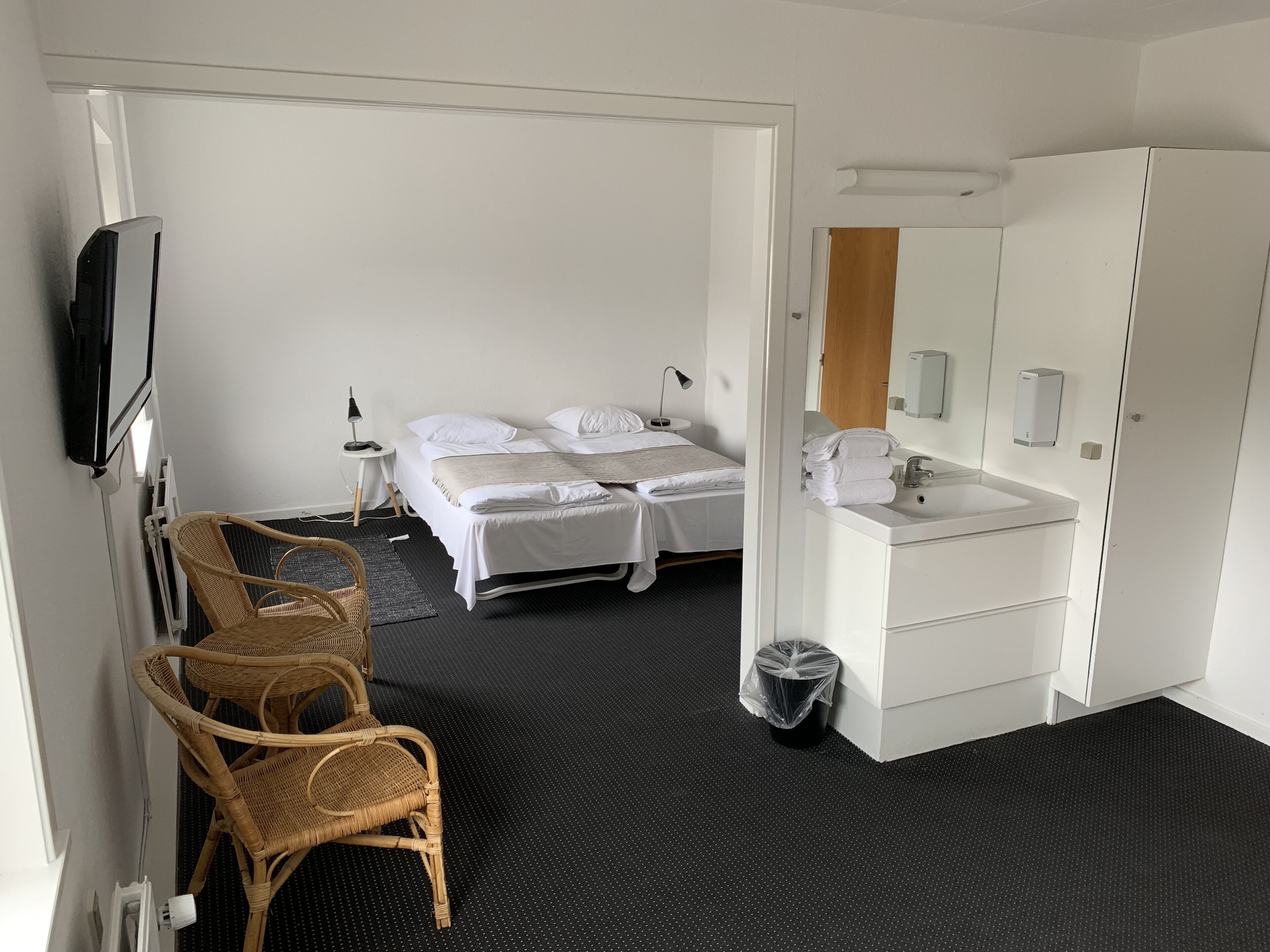 Habitación de hotel con cama, una silla, un escritorio, televisor, baño con lavamanos y espejo, y un cesto de basura.