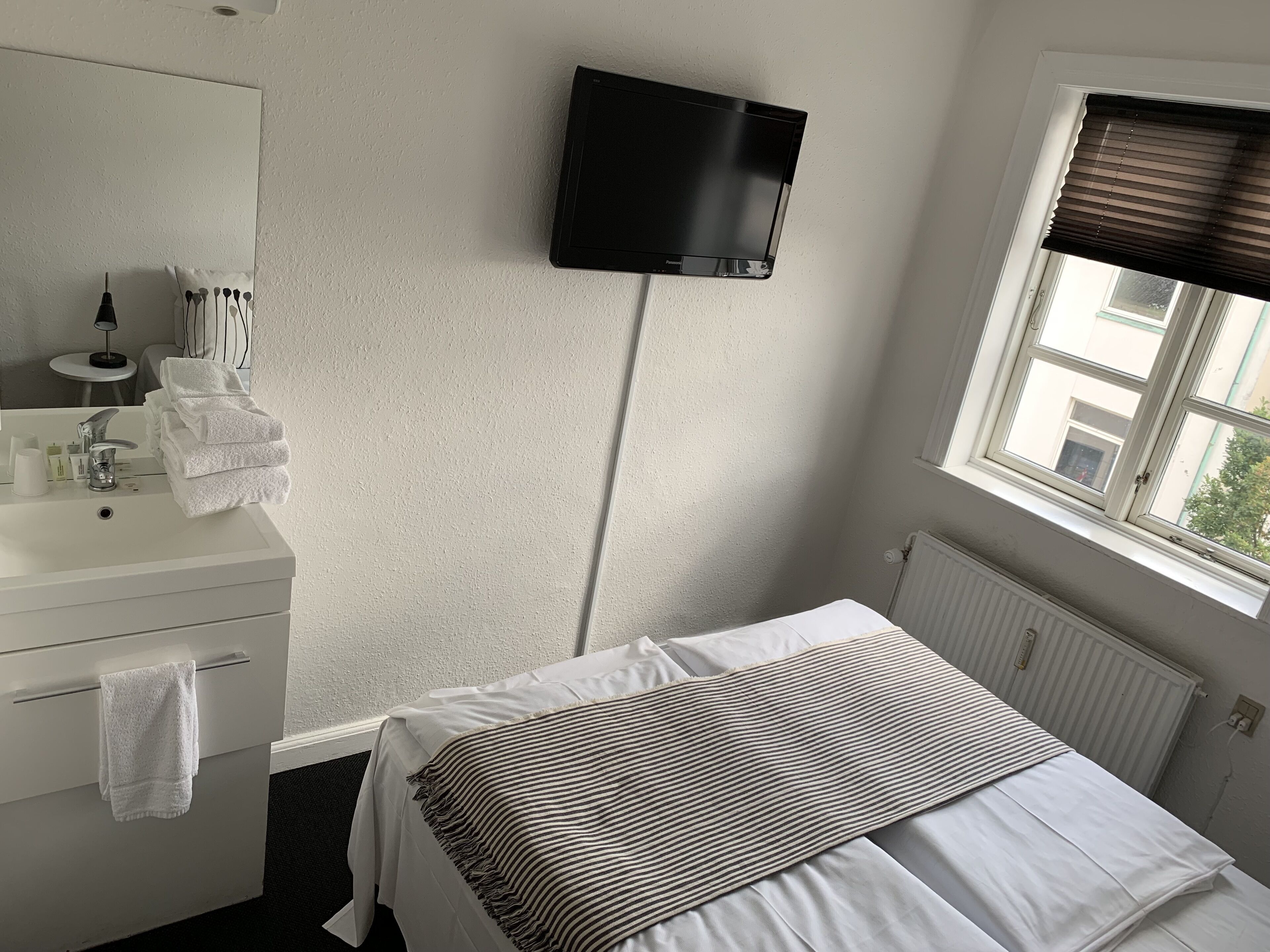 Une chambre d’hôtel avec un lit, un téléviseur fixé au mur, une fenêtre avec des stores, une salle de bain avec un lavabo et des serviettes.