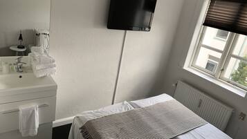 A hotel room with a bed, a TV mounted on the wall, a window with blinds, a bathroom with a sink and towels.