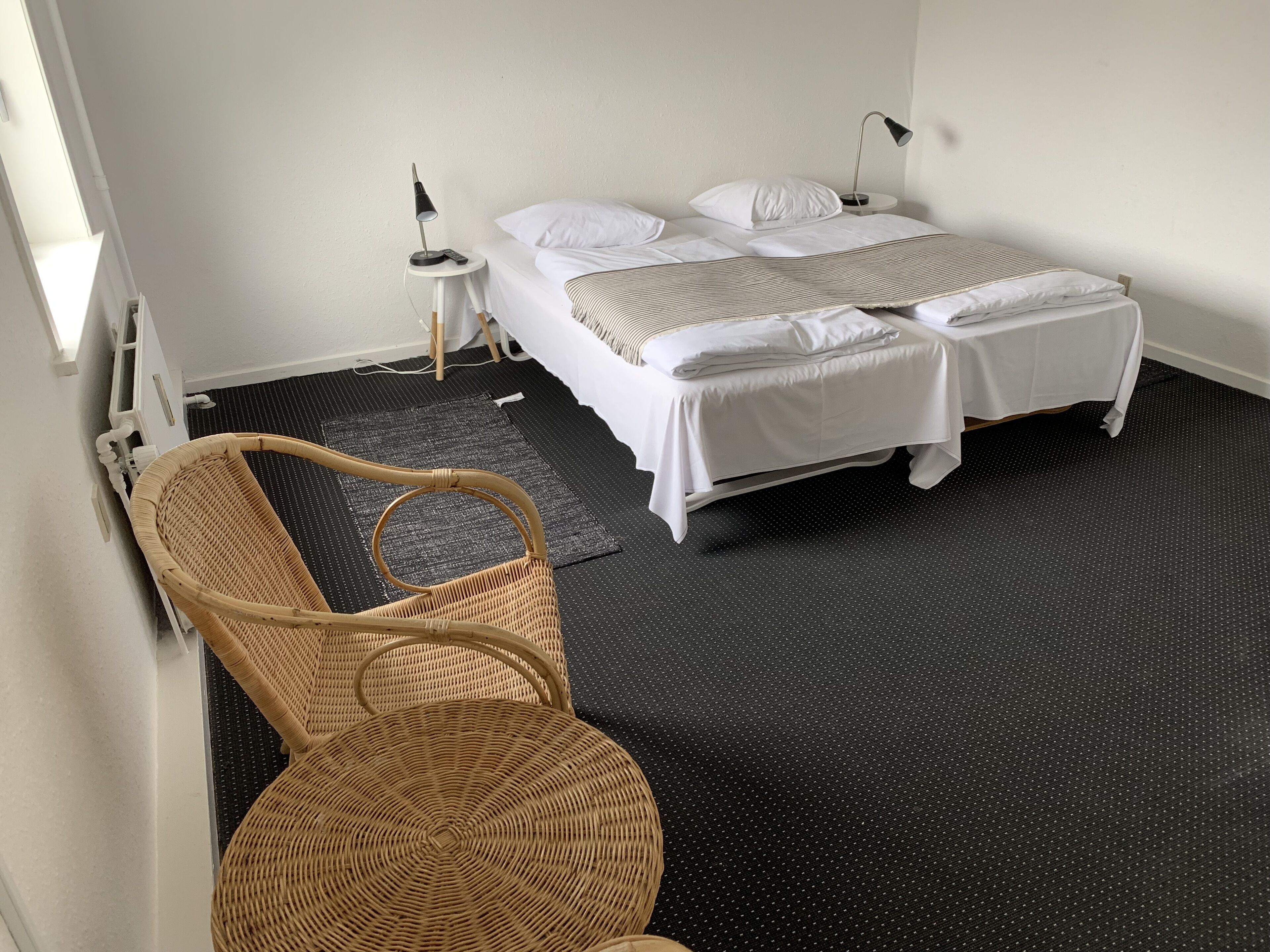A hotel room with a bed, a bedside table with a lamp, a wicker chair, and a black carpet.